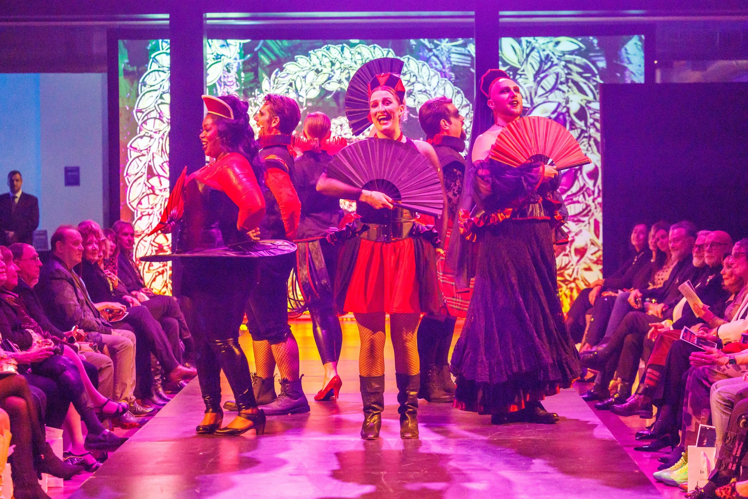 Models walking down the runway at a fashion show, dressed in bold, colorful outfits with accessories, audience sitting on either side, vibrant background projection.