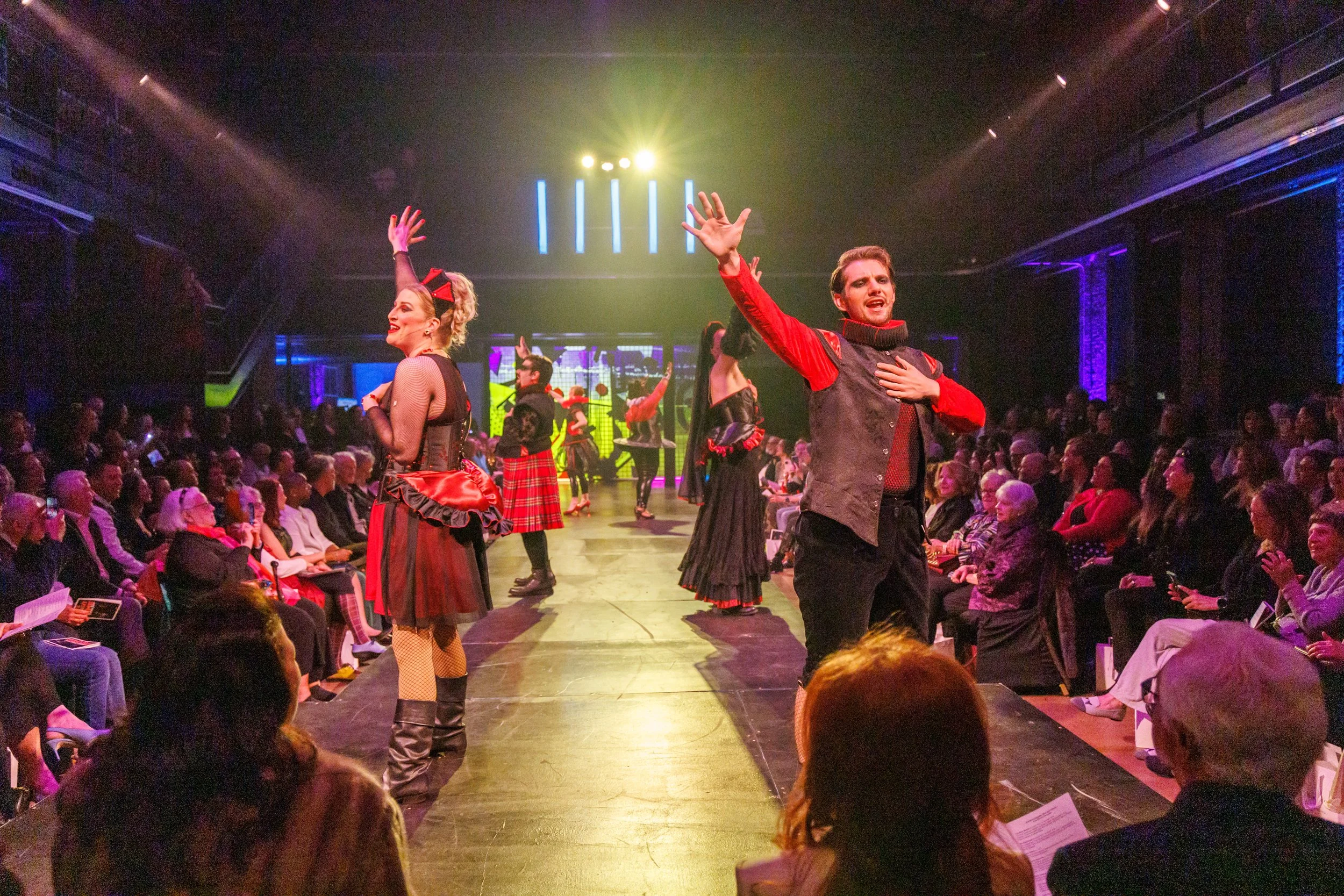People performing a dance or theatrical act on stage in front of an audience. The performers are dressed in colorful costumes, with some in plaid skirts and fishnet stockings, and others in dark vests and bright red accents. The audience is seated, w