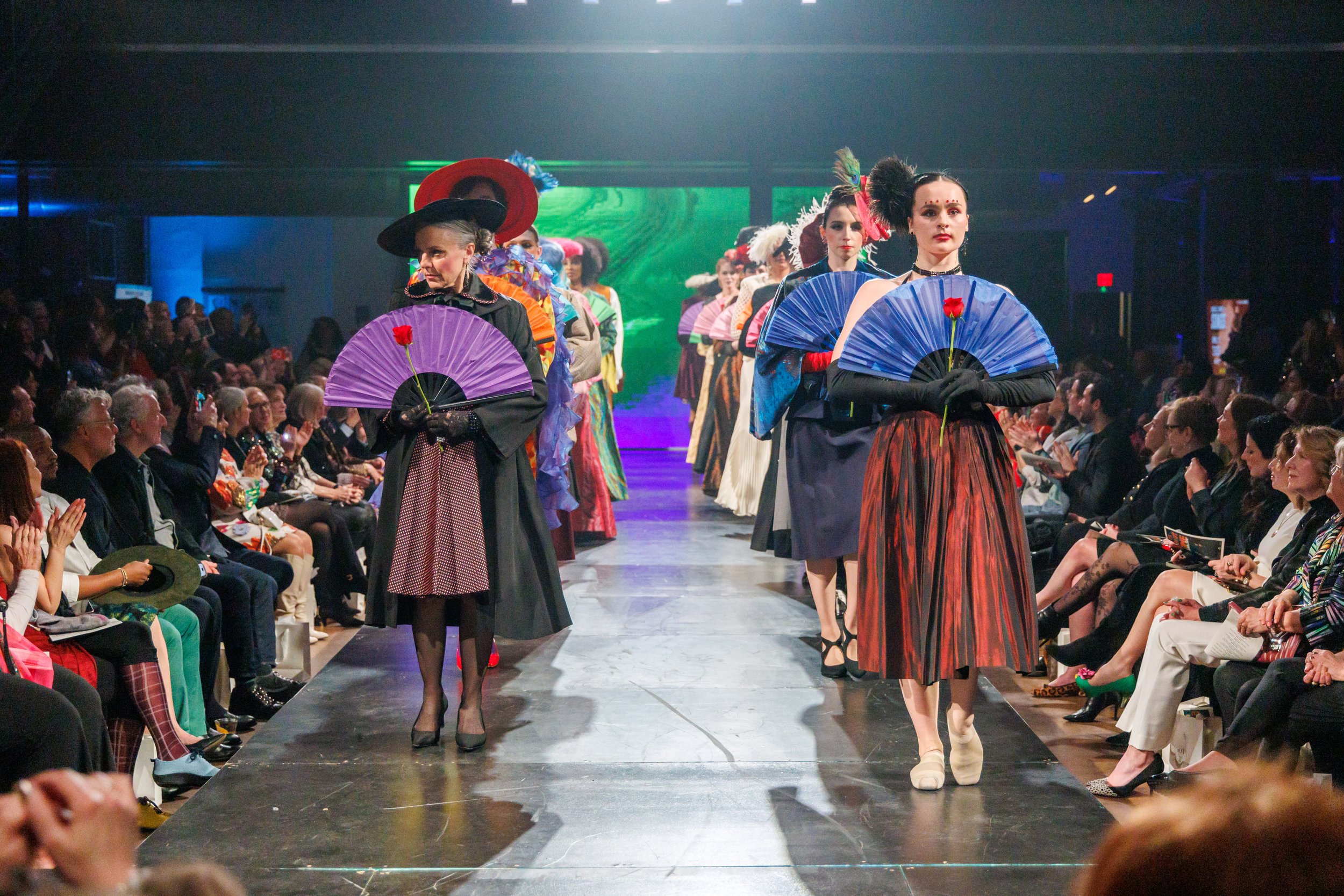 Models walking down a fashion runway during a fashion show, dressed in colorful, vintage-inspired clothing, each holding a paper fan with a red flower. The audience watches and claps on both sides of the runway.