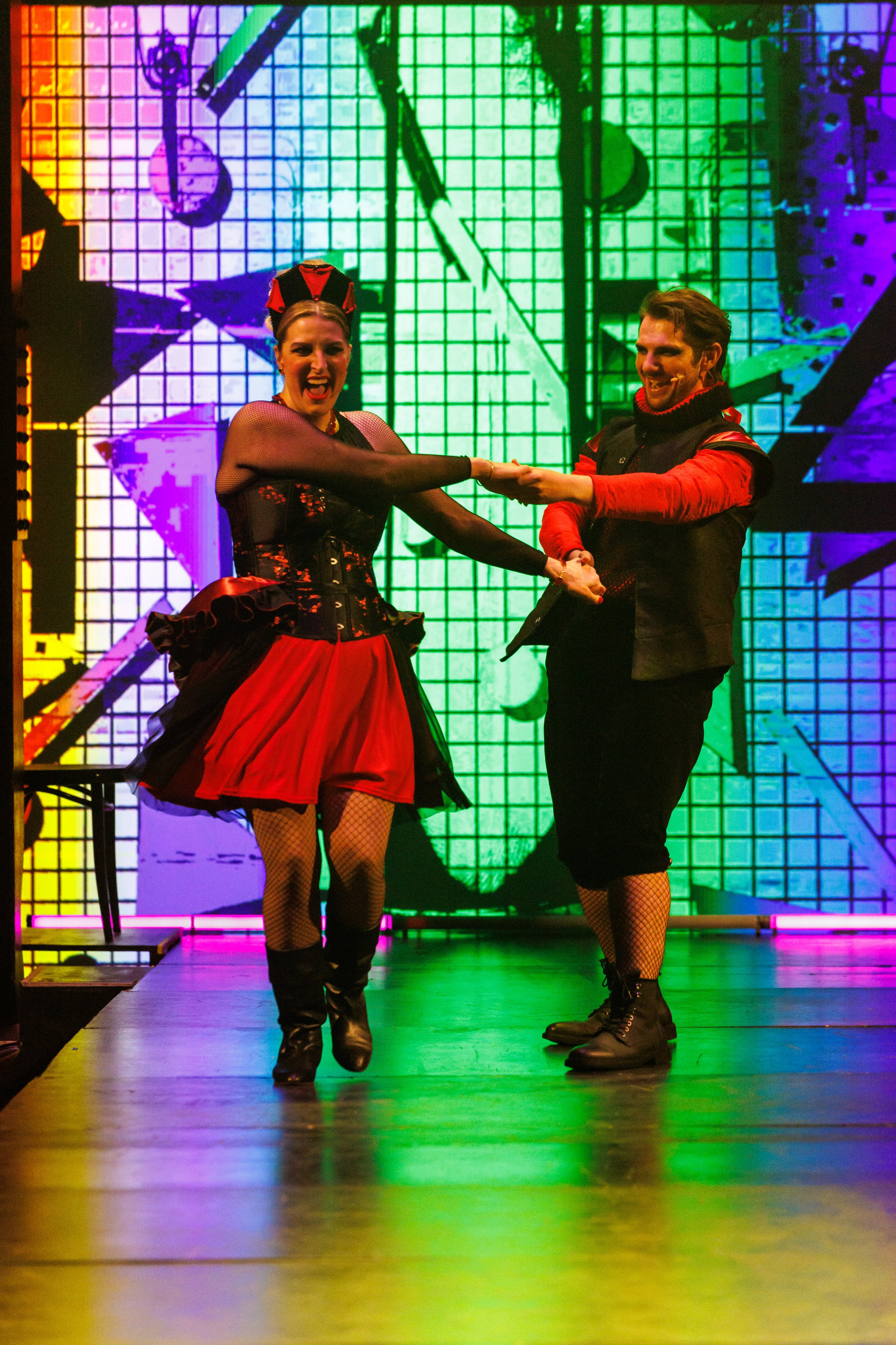 Two performers in colorful costumes dancing or acting on stage with a vibrant, illuminated grid background.
