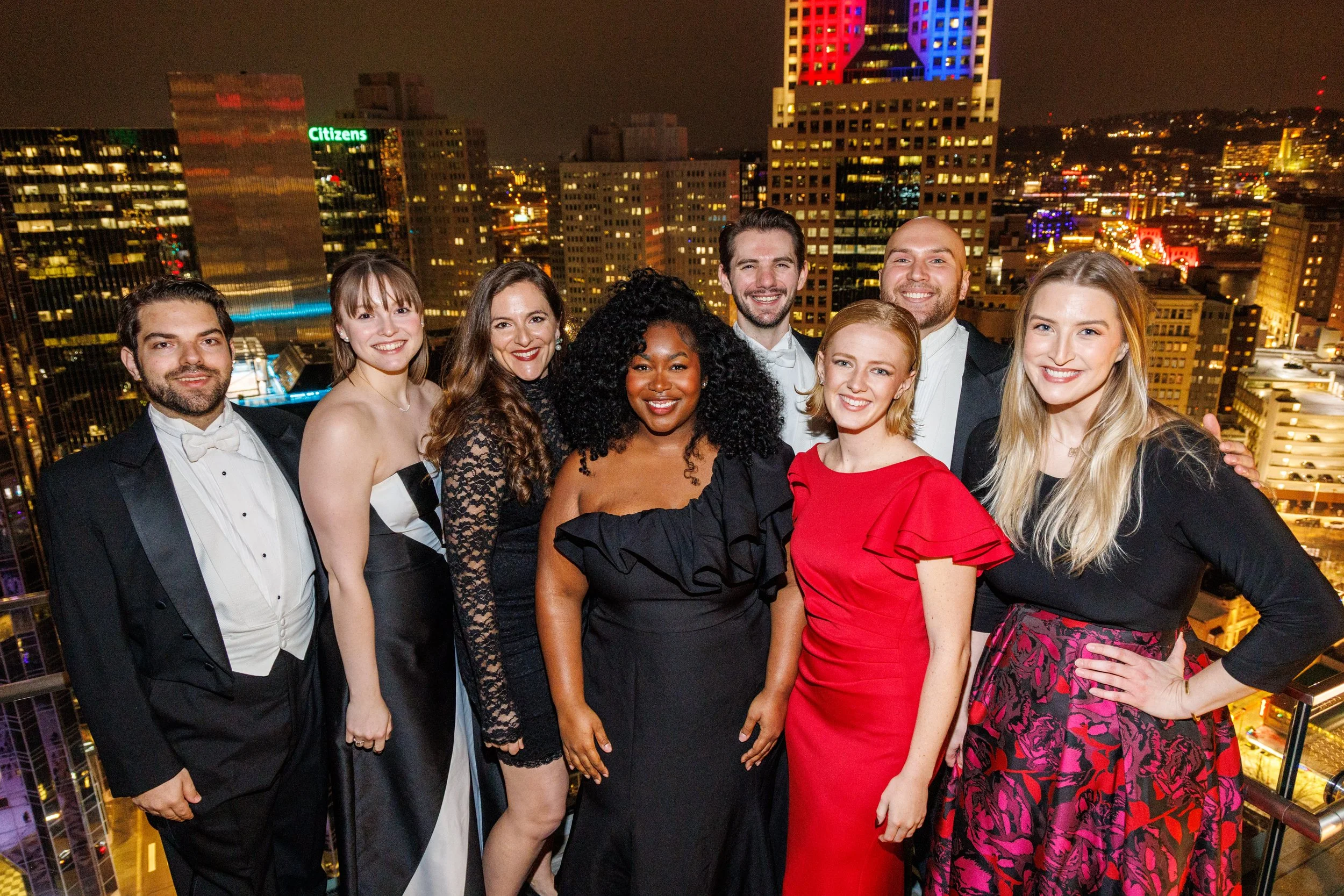 A diverse group of eight well-dressed people smiling on a rooftop at night with a city skyline and illuminated buildings in the background.