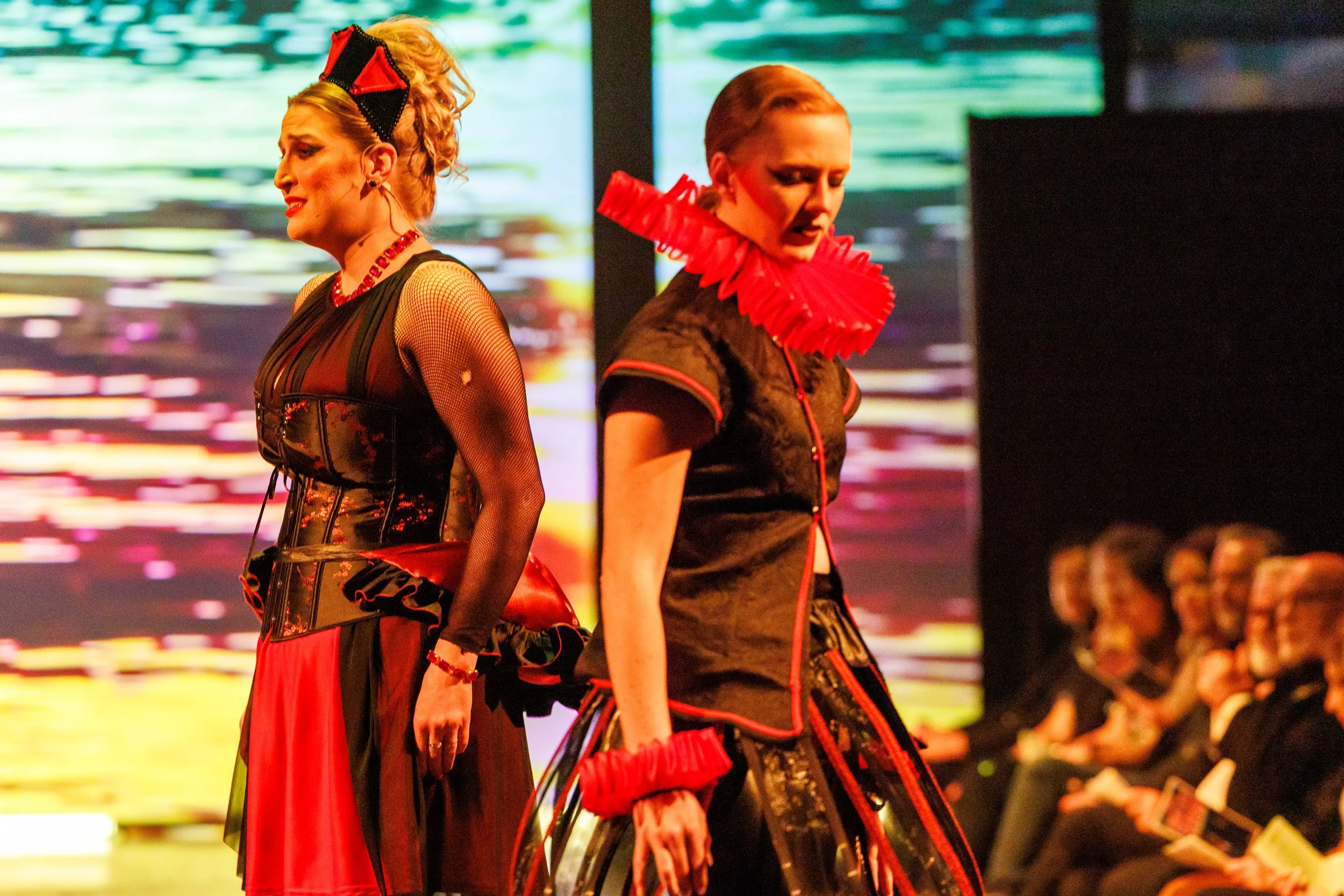 Two women dressed in elaborate black and red outfits walking on a runway during a fashion show, with an audience watching in the background.