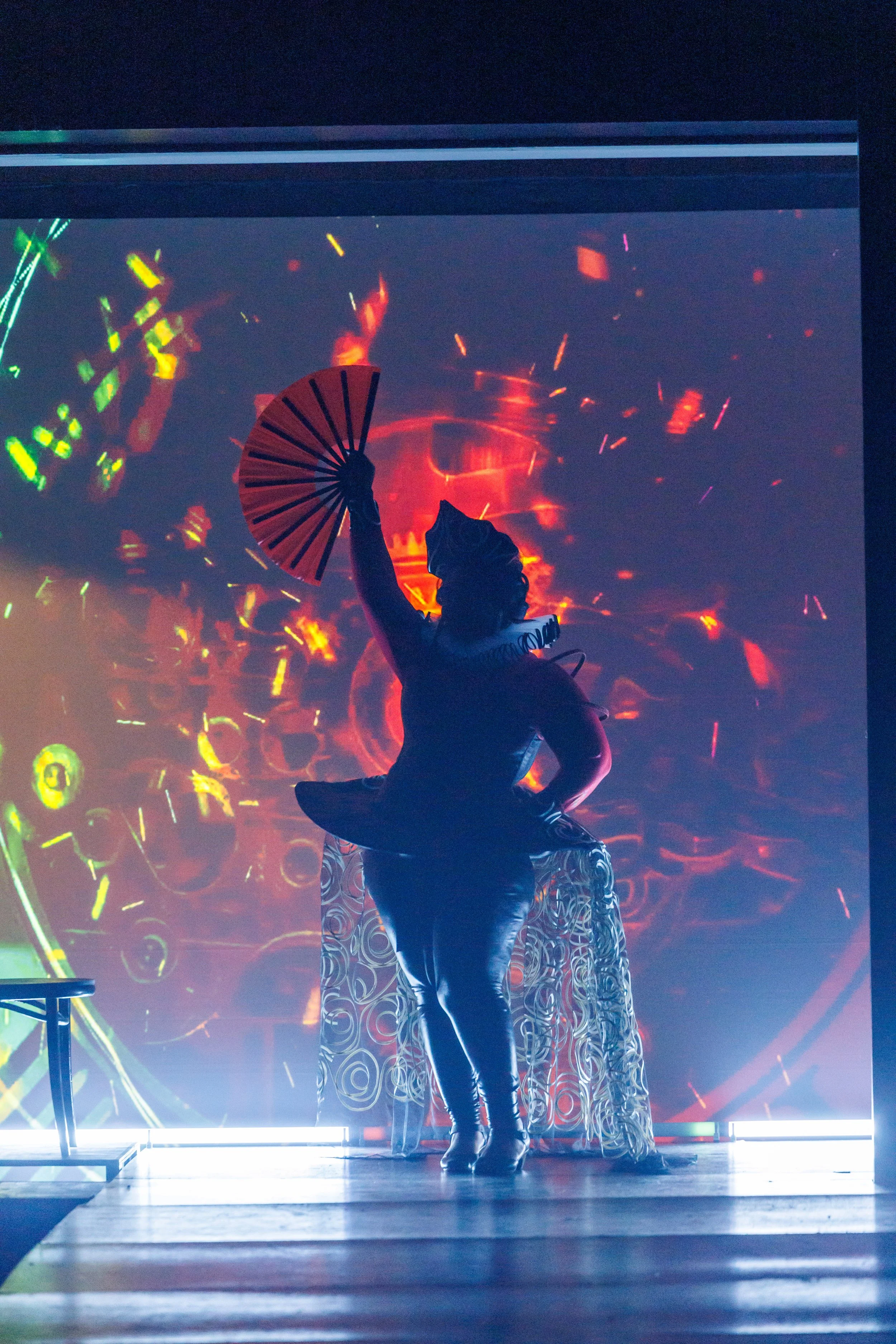 A performer on stage holding a red folding fan with a background of swirling, vibrant, abstract light patterns.