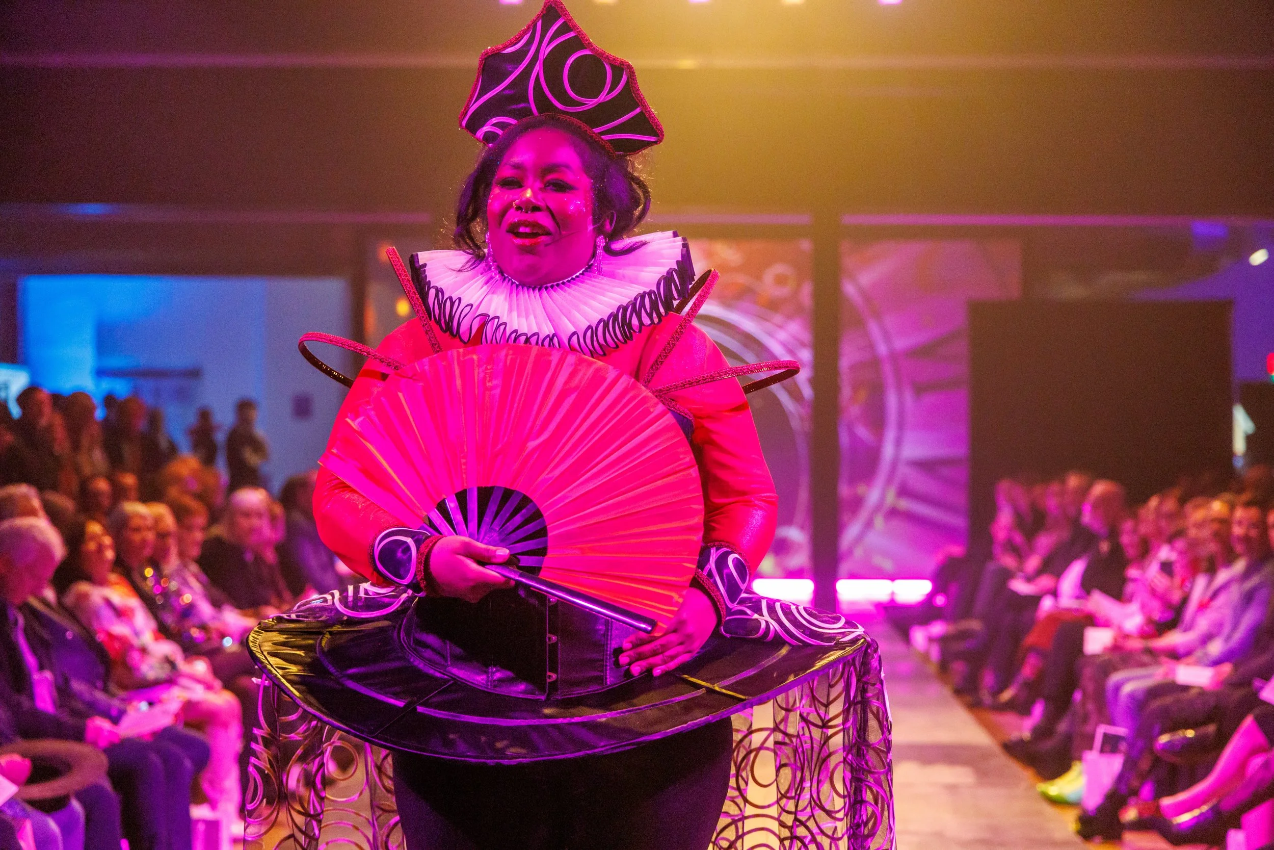 A person dressed in colorful, avant-garde outfit with a large pink fan and elaborate headpiece, walking down a runway in front of an audience at a fashion show.