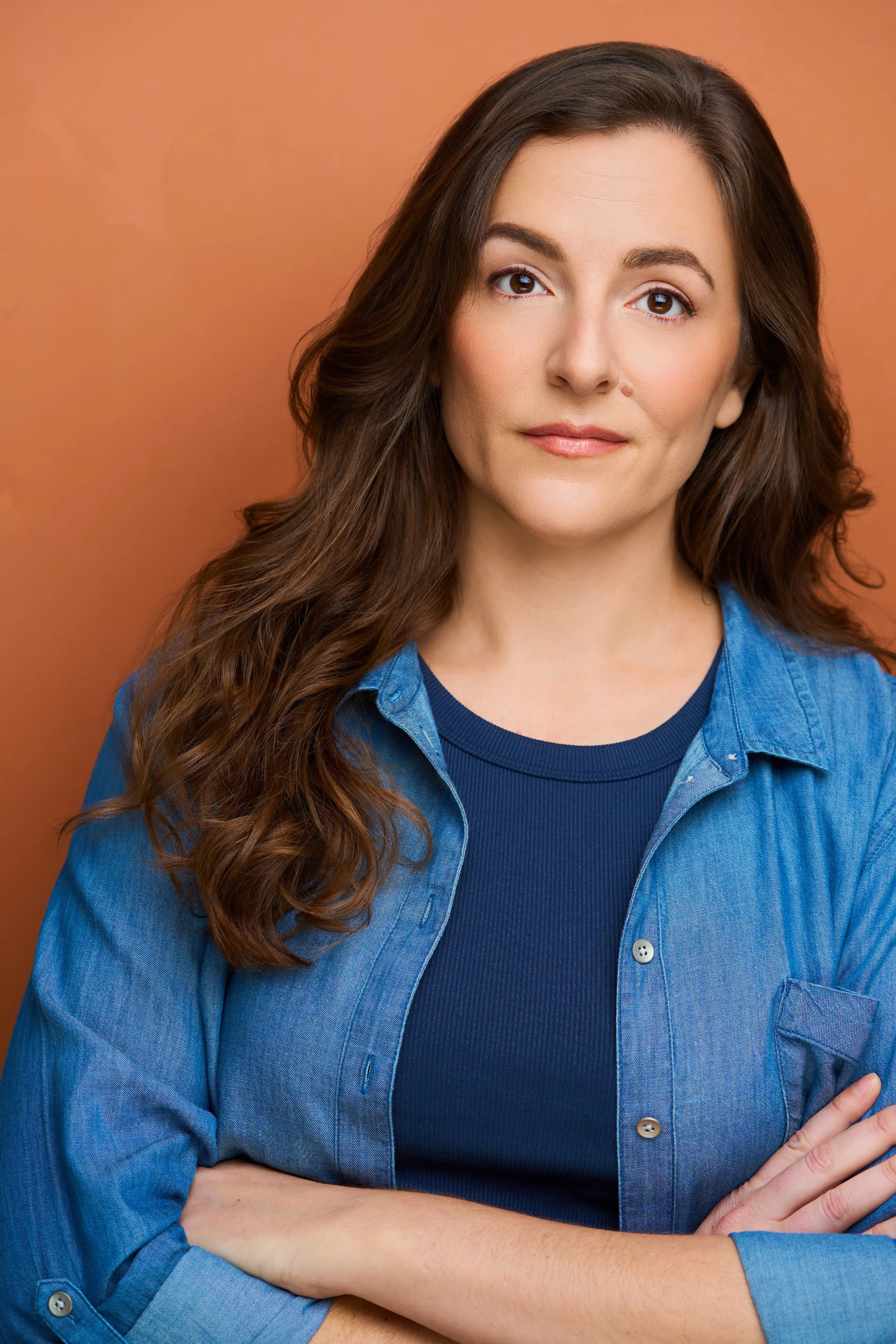 A woman with long brown wavy hair, wearing a blue denim jacket and a navy blue shirt, standing with crossed arms against a warm orange background.