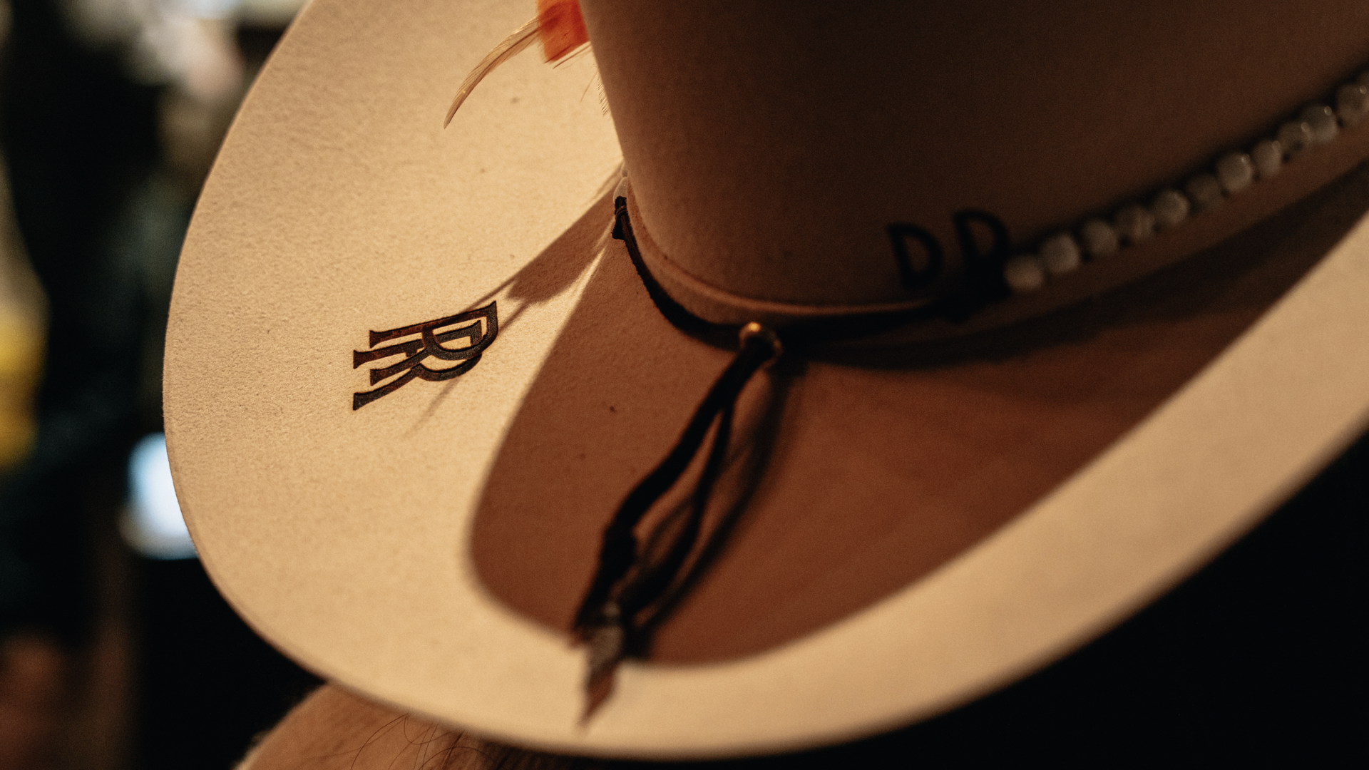 Close-up of a cowboy hat with the letter 'RR' visible on the brim, a leather band with decorative beads, and a faint letter 'D' on the crown.