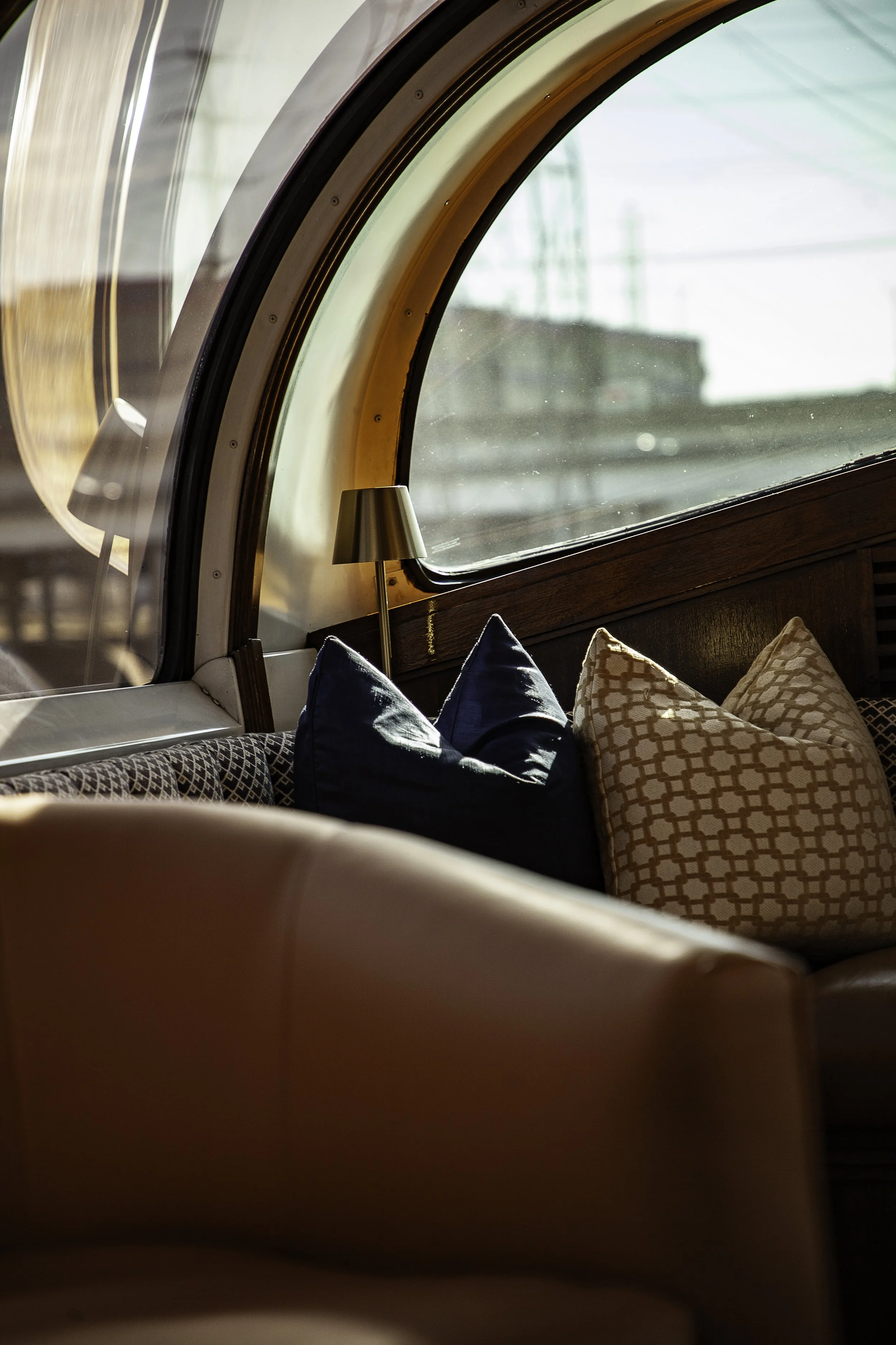 Inside a modern train cabin showing a curved window, a small lamp, and cushioned seating with patterned pillows.