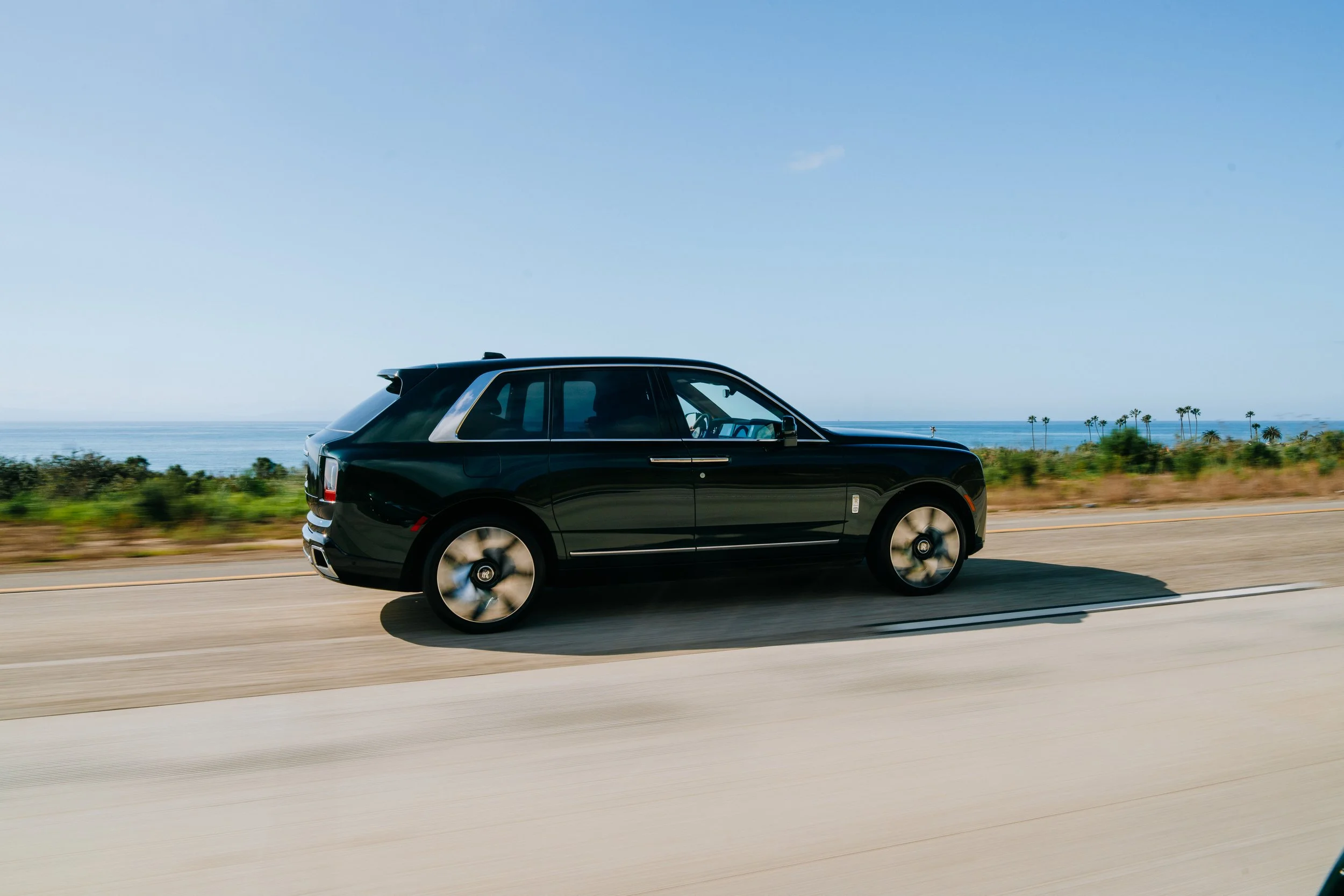 A black luxury SUV driving on a coastal highway with palm trees and the ocean in the background under a clear blue sky.