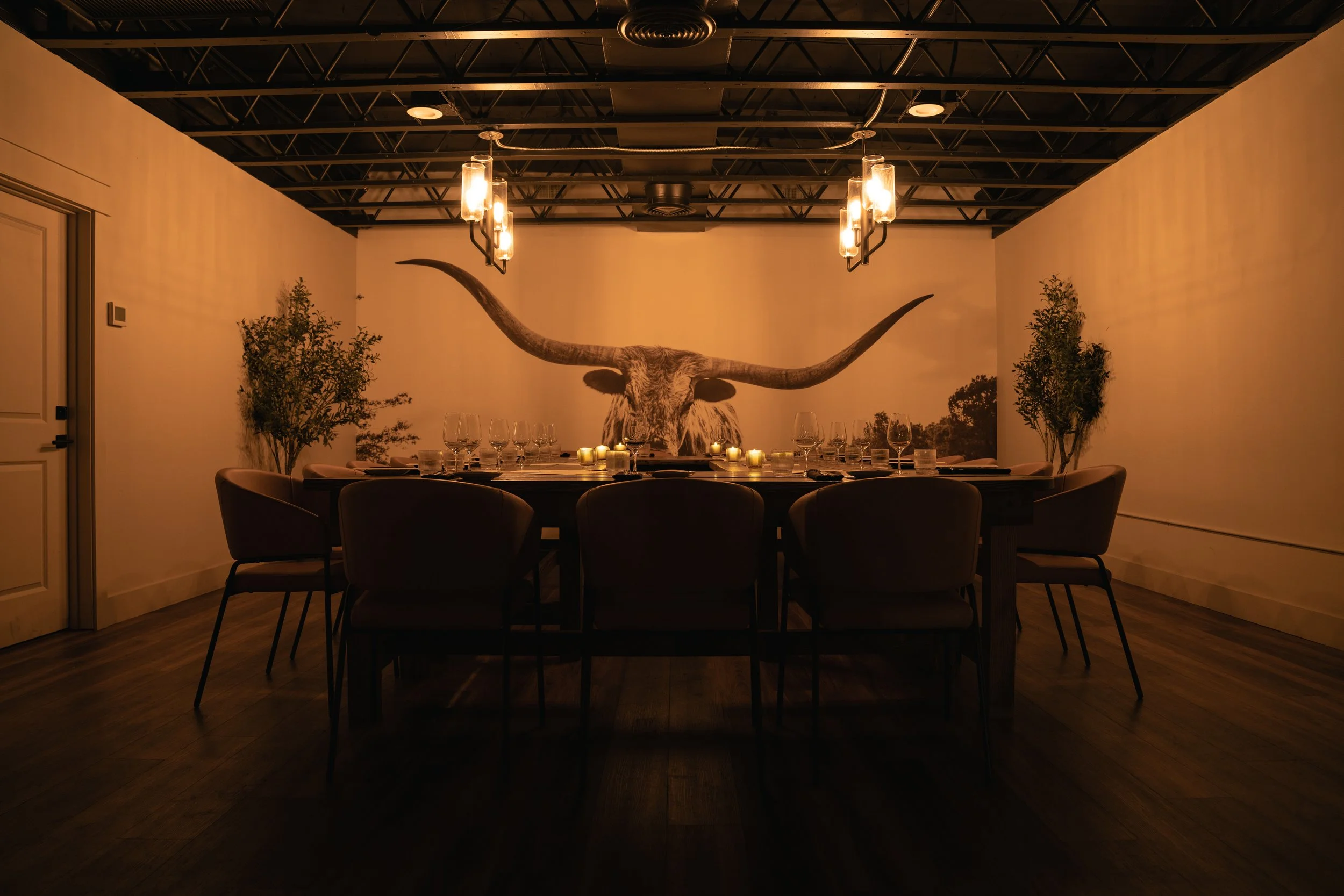 A dining room with a long table set with glasses, plates, and candles. A large mural of a buffalo with long horns decorates the wall behind the table. The room has a modern industrial ceiling with hanging lights and two potted plants on each side of the mural.