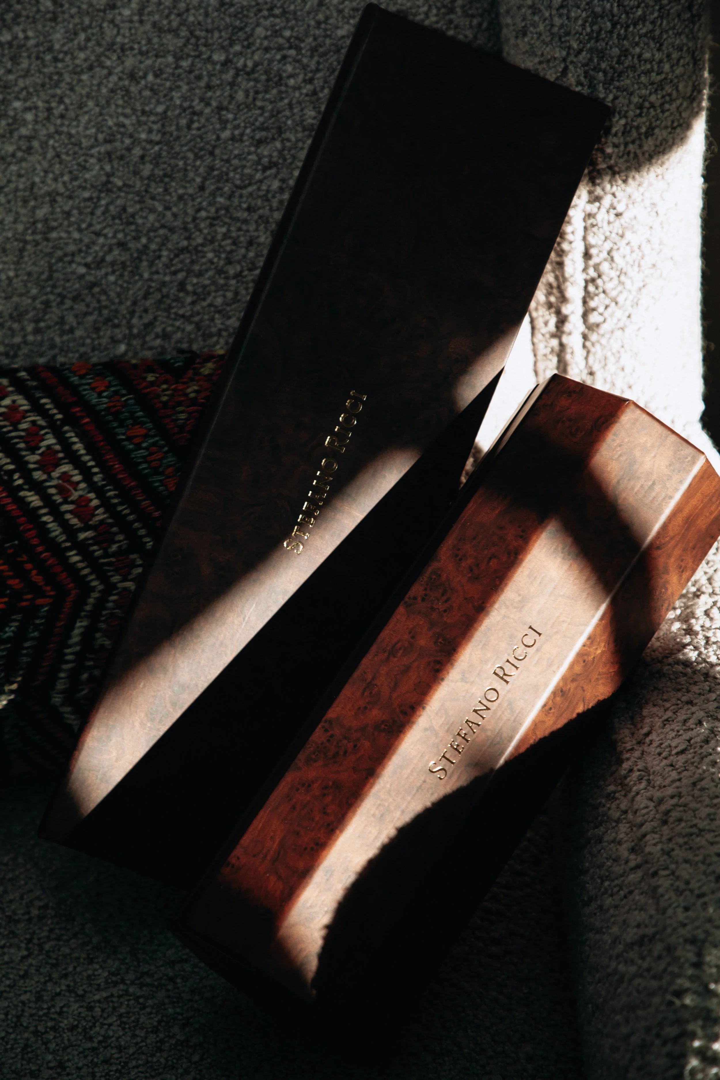 Close-up of two designer handbags, one black and one brown, on a textured dark surface. The bags display the brand name 'STEANO RICCI' in gold lettering.