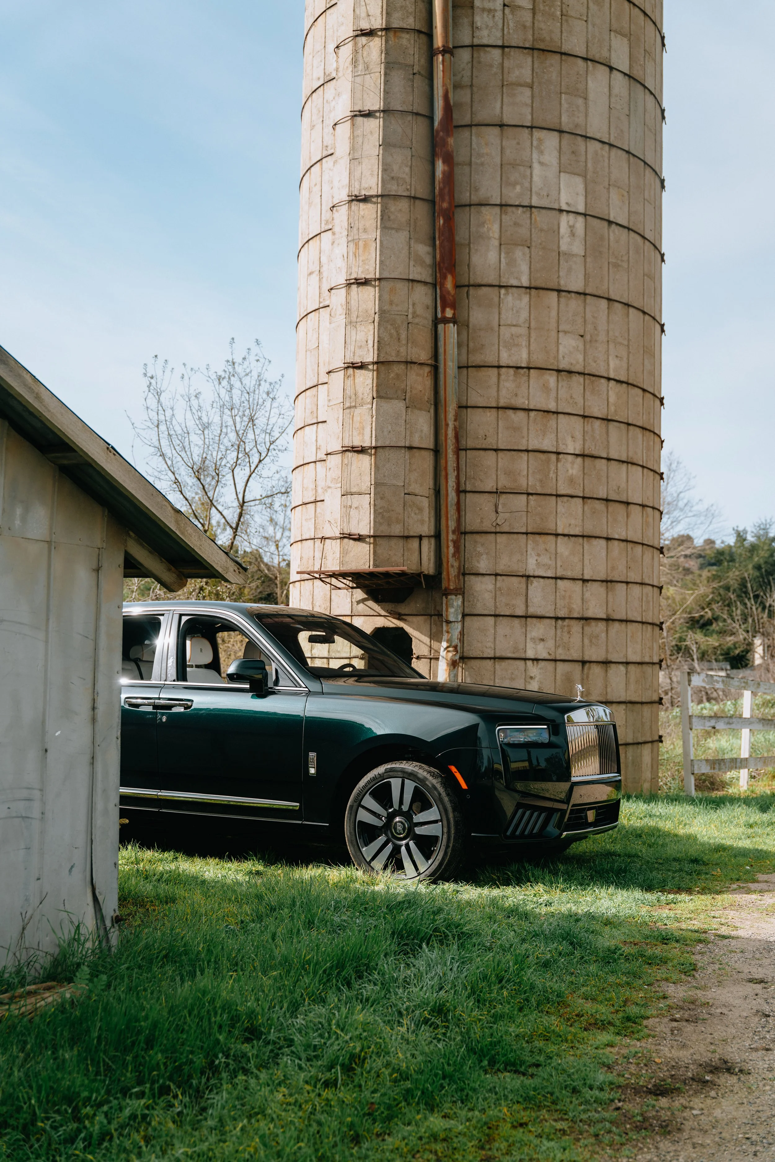 A dark green luxury car parked on a grassy area beside a building with a large, cylindrical, weathered silo in the background. The scene is outdoors with clear skies and some trees visible in the distance.