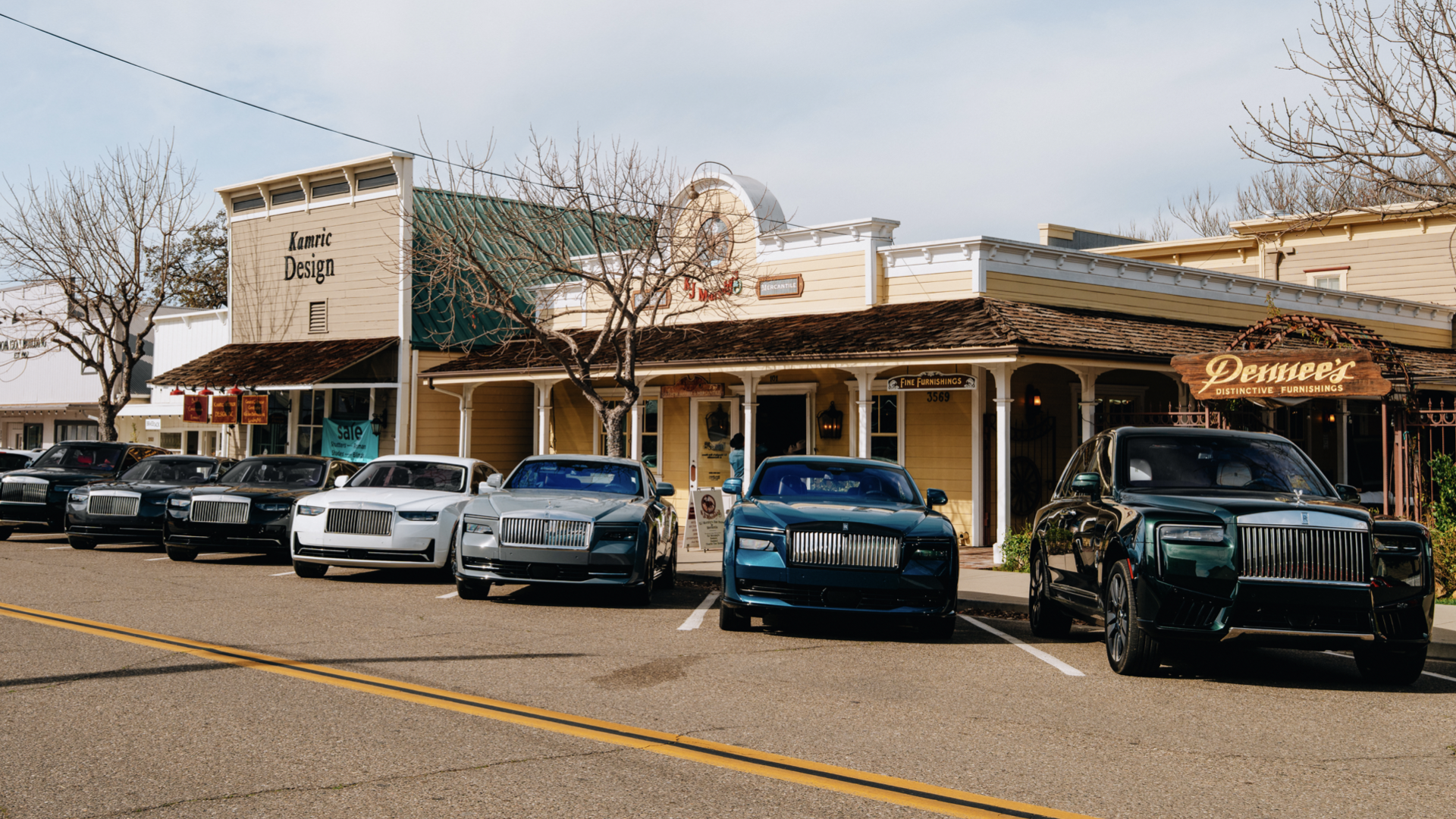 Street view of a row of Rolls-Royce cars parked in front of small shops, including a furniture store and an antique shop, in a small town with bare trees and a cloudy sky.