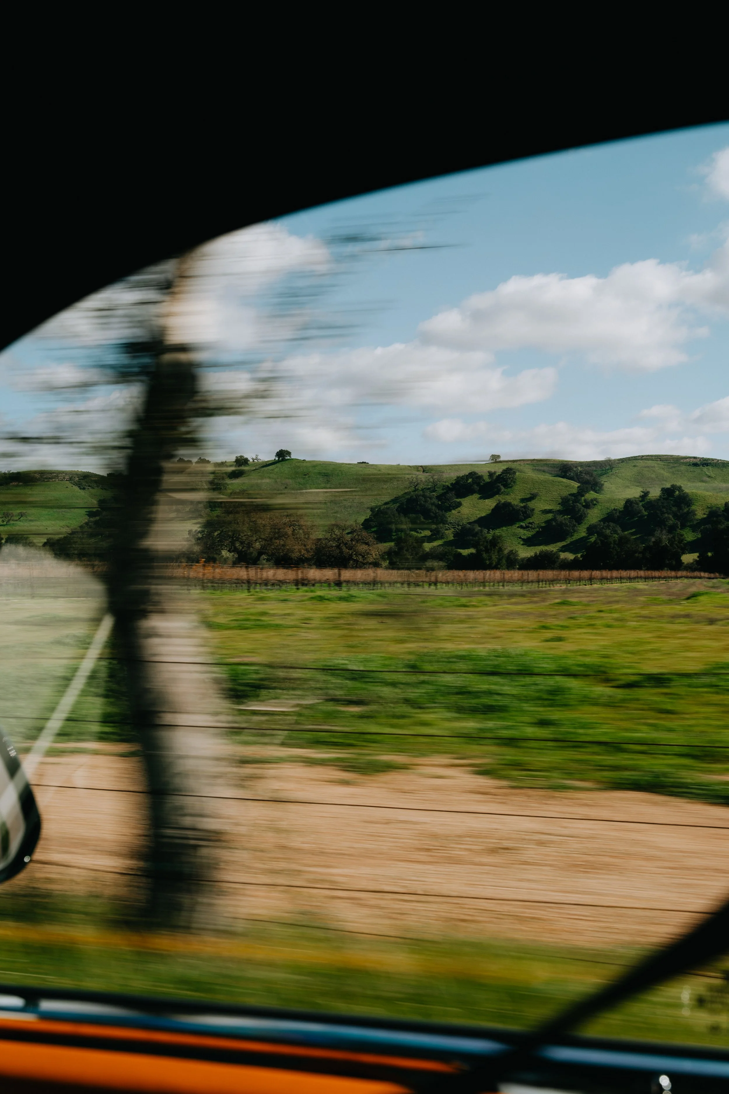 Blurred landscape of green rolling hills under blue sky with scattered white clouds, viewed from inside a moving vehicle with a black interior and part of the dashboard visible.