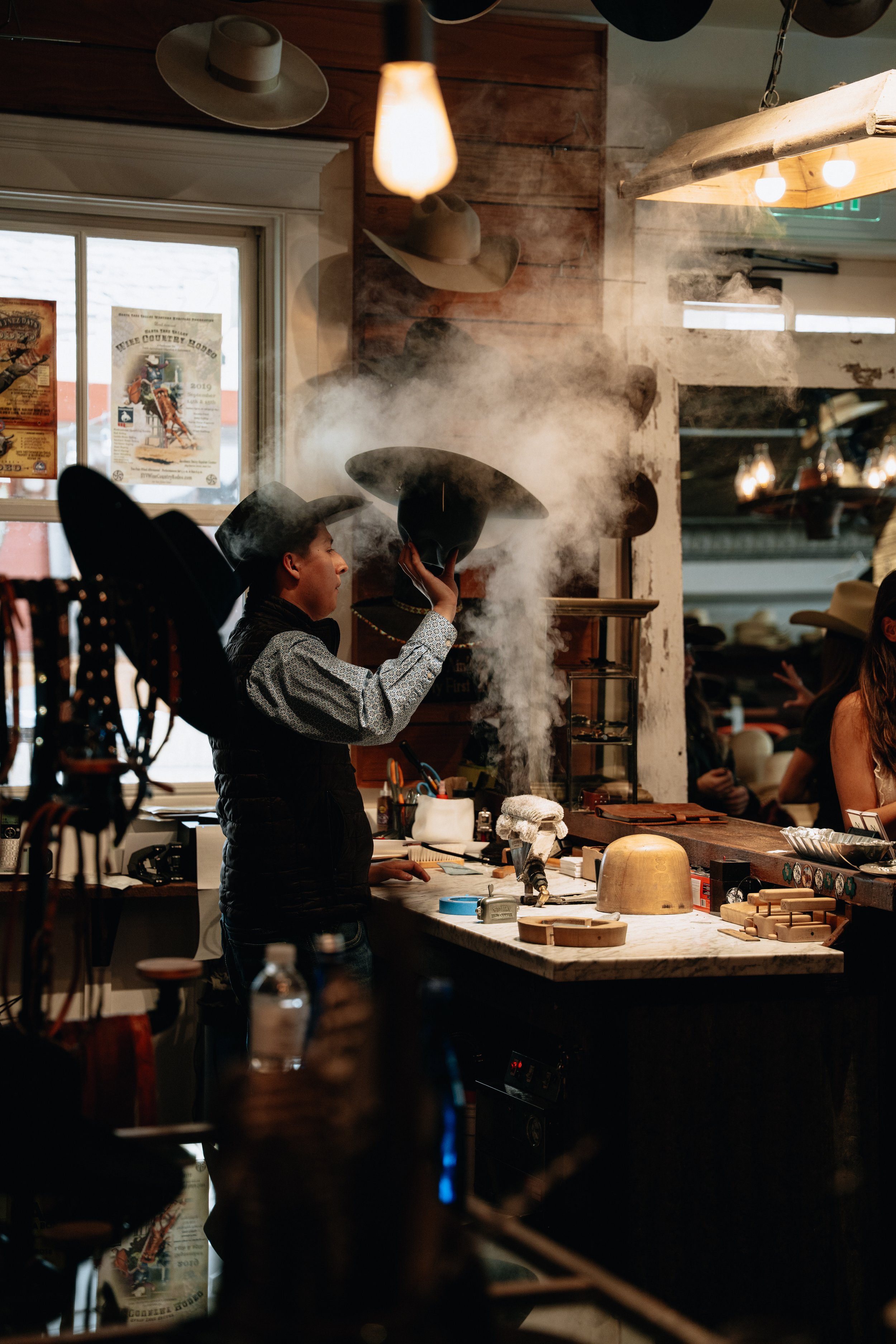 A cowboy wearing a cowboy hat, working in a rustic workshop with various tools and wooden objects on the counter, and steam rising from a bowl or container.