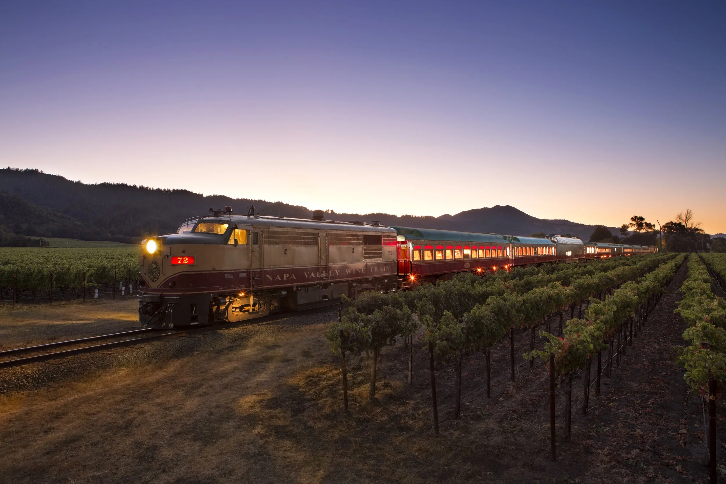 A vintage Napa Valley wine train traveling through vineyards at dusk with mountains in the background.