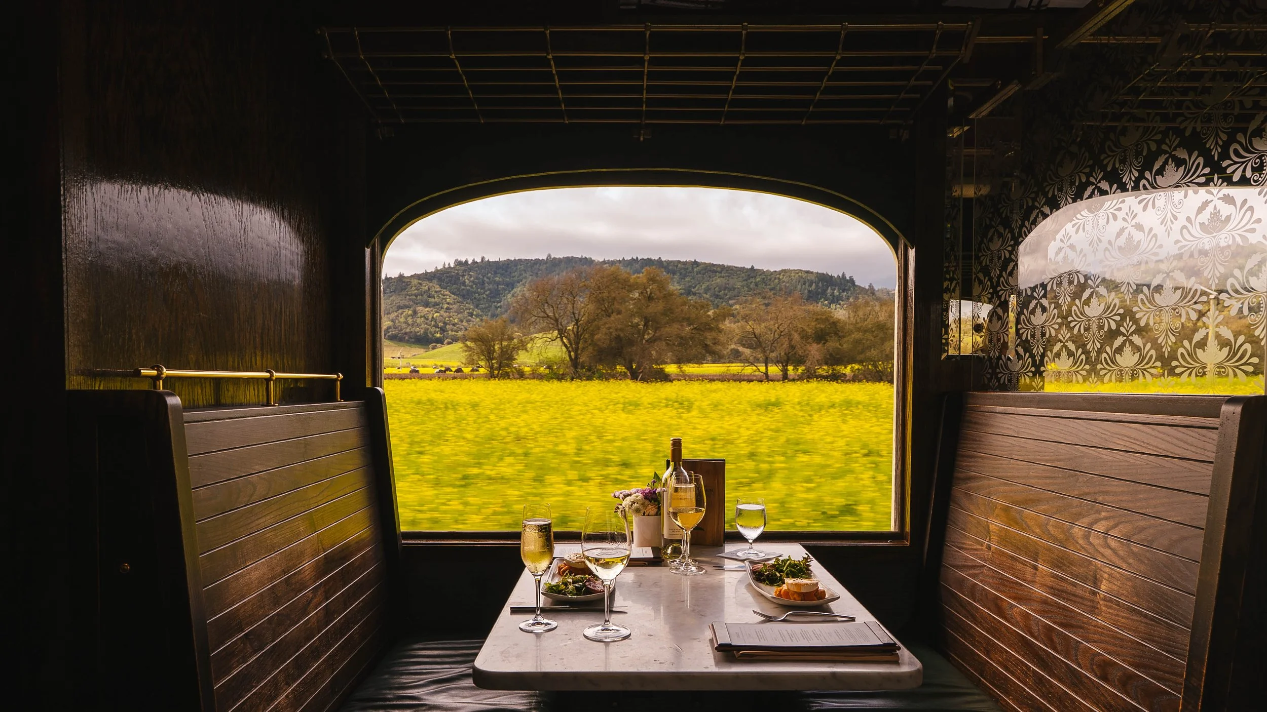 View of a dining table inside a train with large windows revealing a lush green landscape and rolling hills outside, with some trees and clouds in the sky.