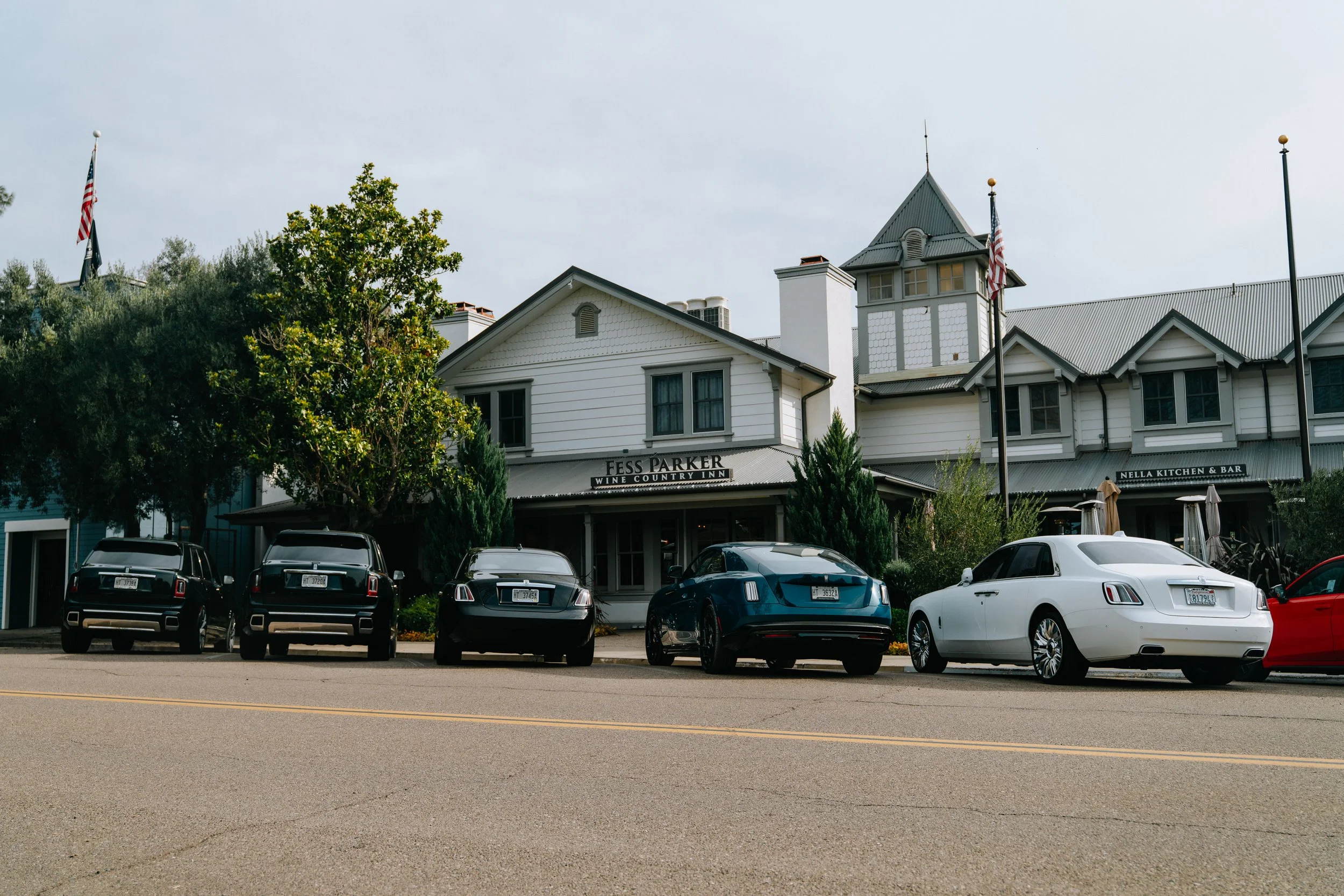 Parking lot in front of a white building with black signs, trees, and a cloudy sky. Several parked cars are visible, including a black SUV, a dark gray Mercedes, and a white car. Flags are flying, and the building has a sign reading 'Fess Parker Wine Country Inn' and 'Nella Kitchen & Bar'.