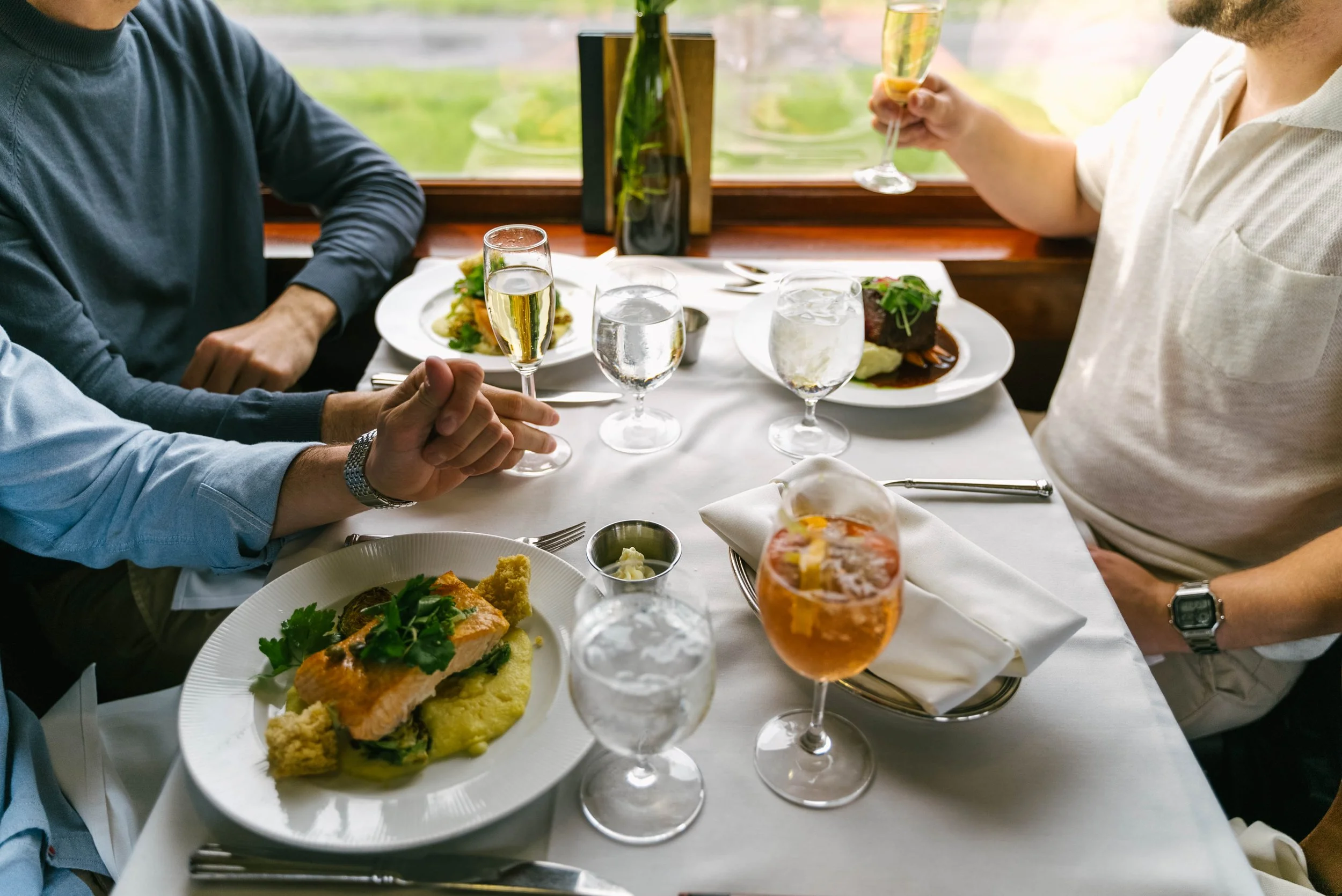 People dining at a restaurant table with various dishes including salmon, vegetables, and drinks like champagne and water.