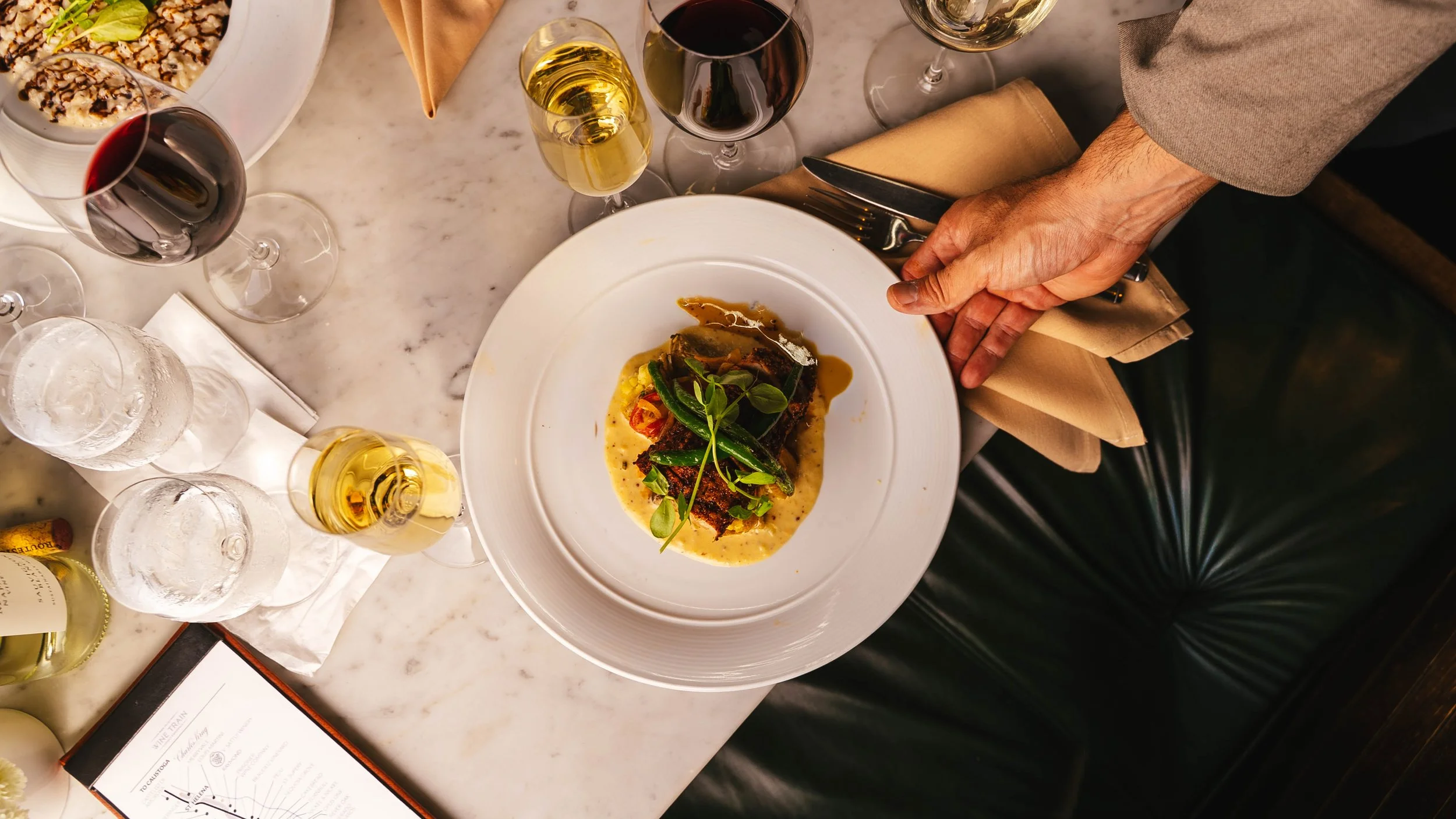A top-down view of a dinner plate with a fish entree garnished with greens and vegetables, on a marble table. Surrounding the plate are glasses of red and white wine and water, as well as a folded napkin and cutlery. Part of a salad and wine bottle are also visible.