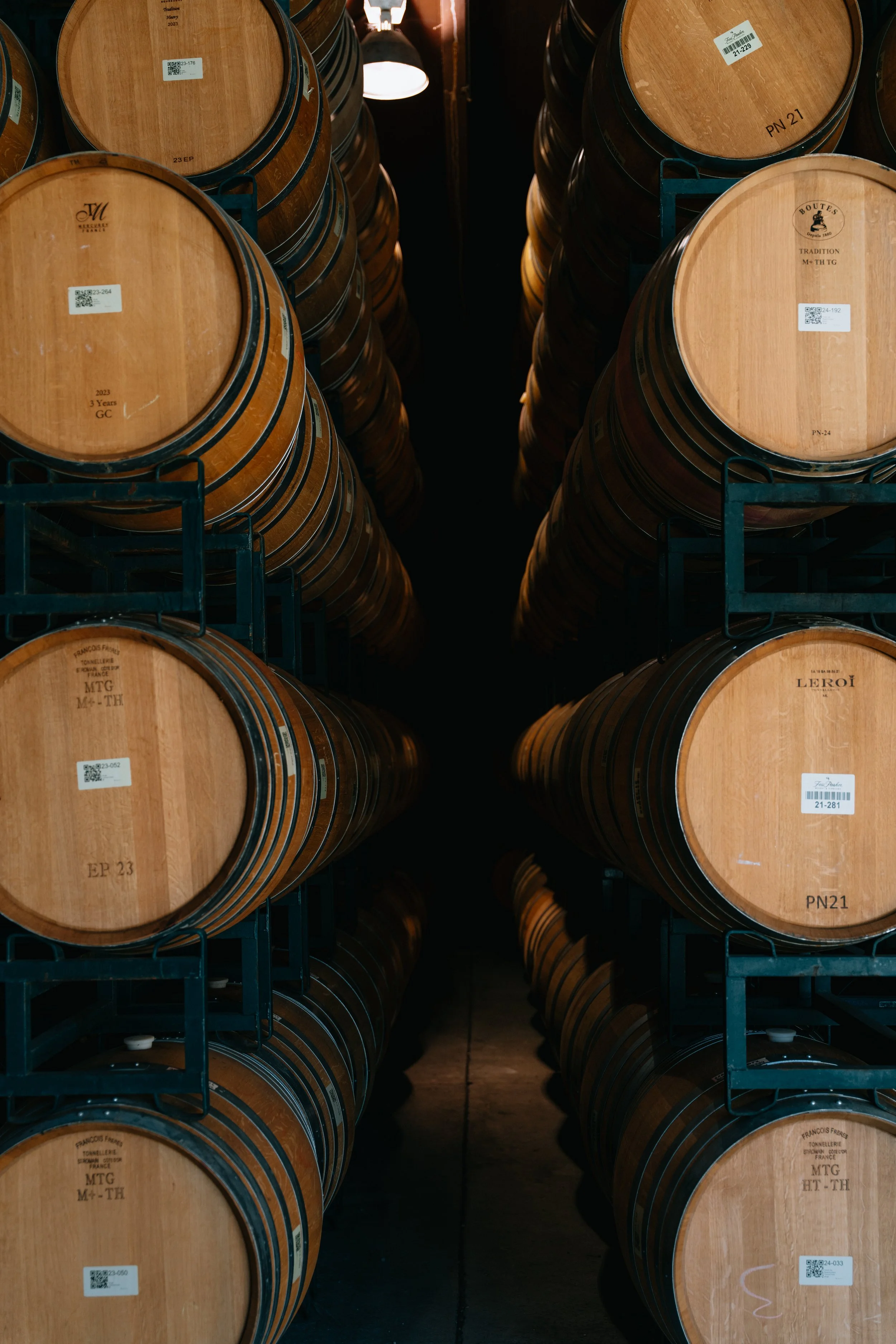 Rows of wooden barrels stacked in a storage area, with some barrels bearing labels and QR codes, under dim lighting.