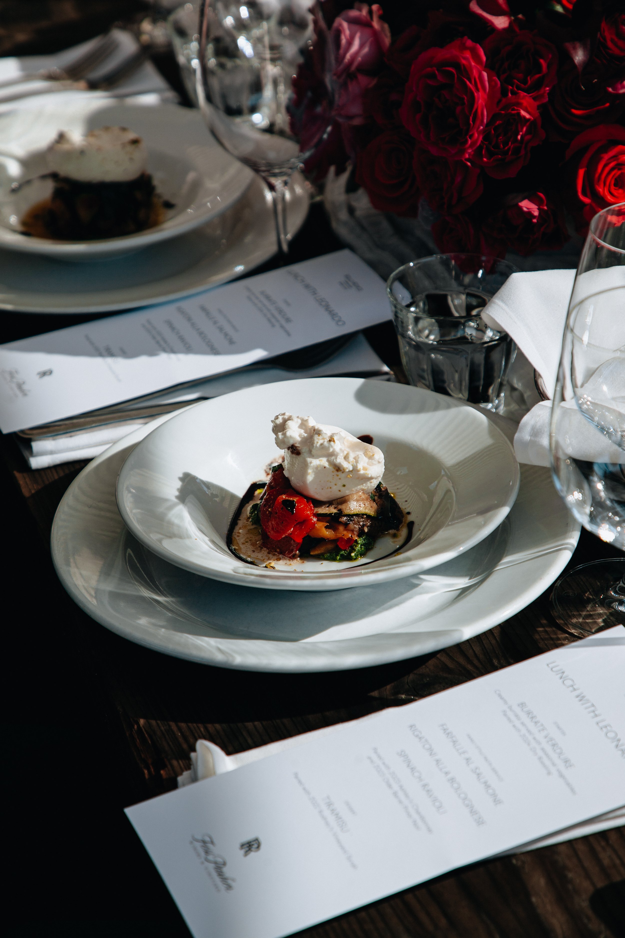 A dining table with a white plate holding a dish topped with a scoop of ice cream, a small bowl of dark sauce, and a cherry. There is a bouquet of red roses in the background, wine glasses, and printed menus or programs on the table.