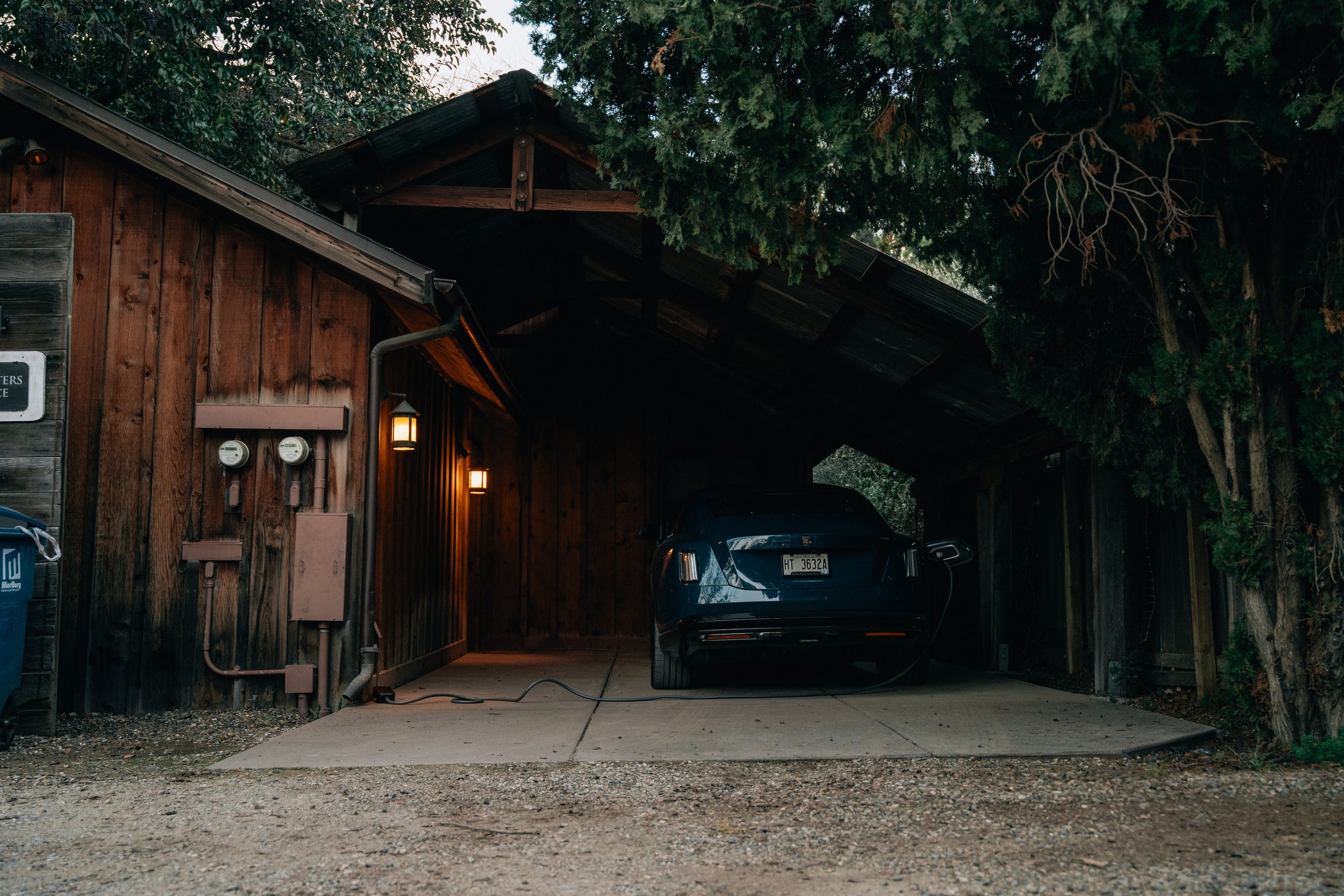 A wooden house with a car parked in the driveway, surrounded by trees with dense foliage. The house has outdoor lights turned on, electrical meters on the wall, and a power cord on the ground.