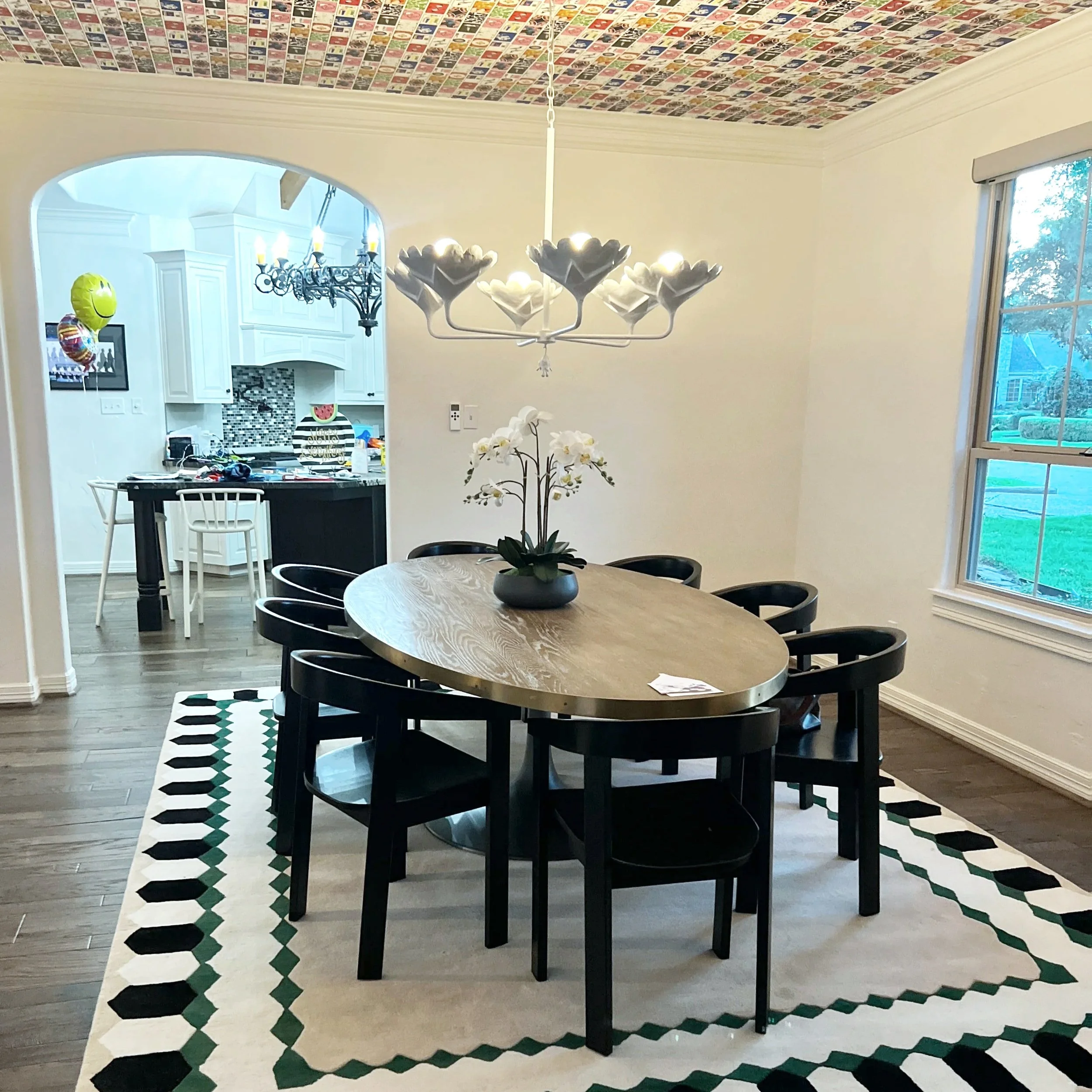 A dining room with a round wooden table and black chairs, a white orchid centerpiece, a geometric patterned rug, a window to the right, and a chandelier with flower-shaped lights above. The ceiling is decorated with colorful small pictures.