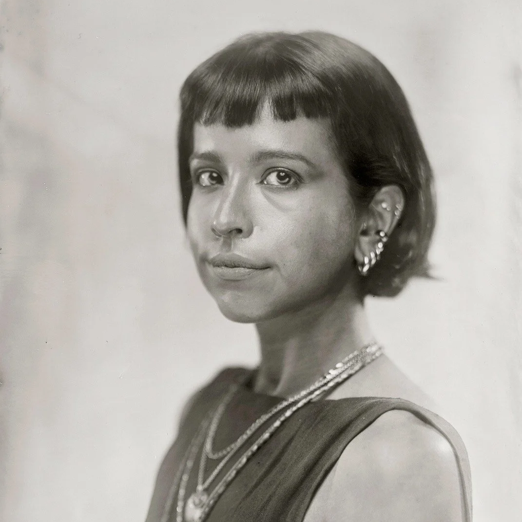 Black and white portrait of a woman with short dark hair, wearing earrings and layered necklaces, looking at the camera.