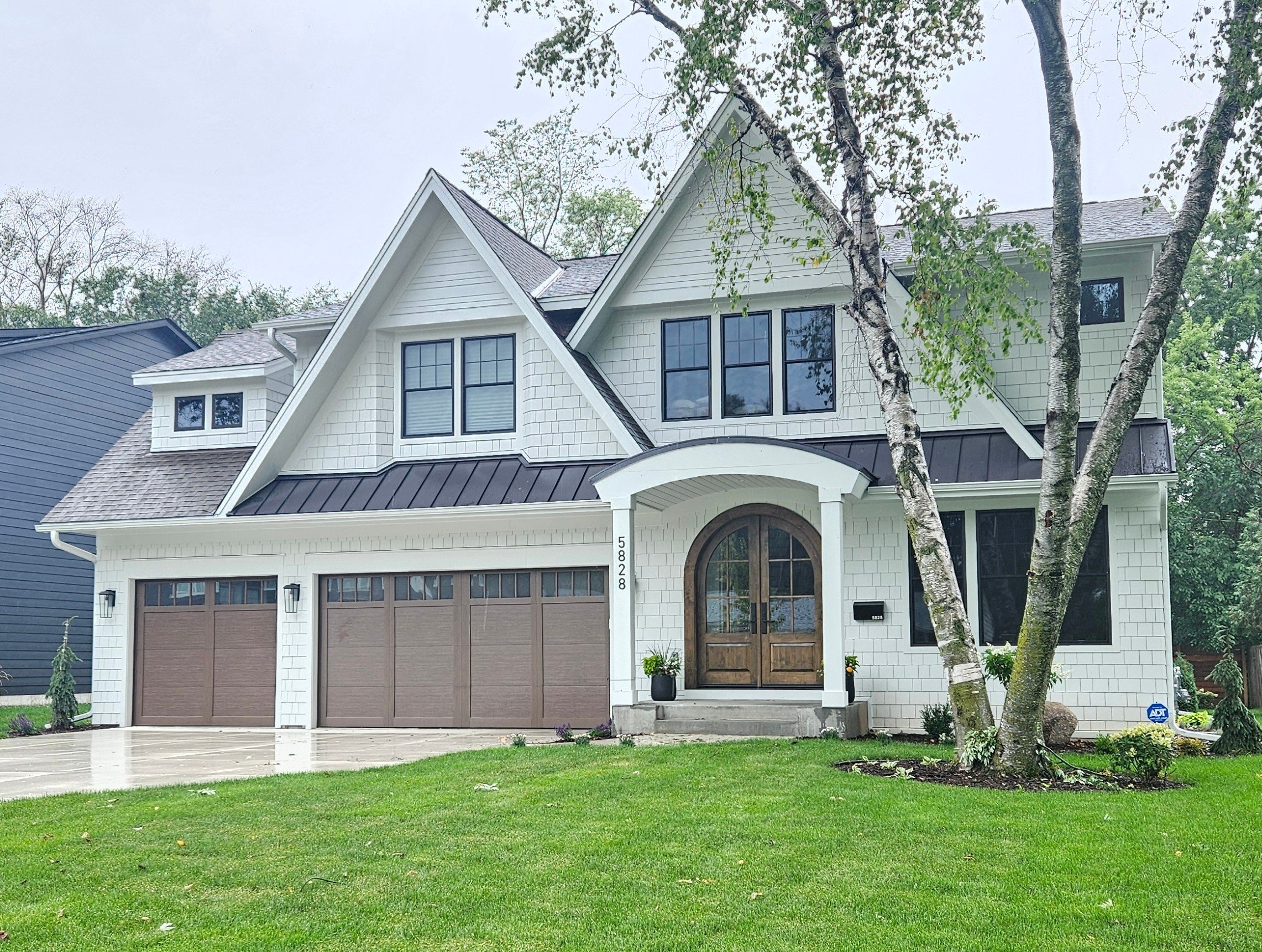 Modern Shingle Style cottage exterior with white shakes, arched double entry doors, dark metal accent roof, black grid windows, and three-car wood carriage garage doors at 5828 Kellogg Ave S, Edina MN