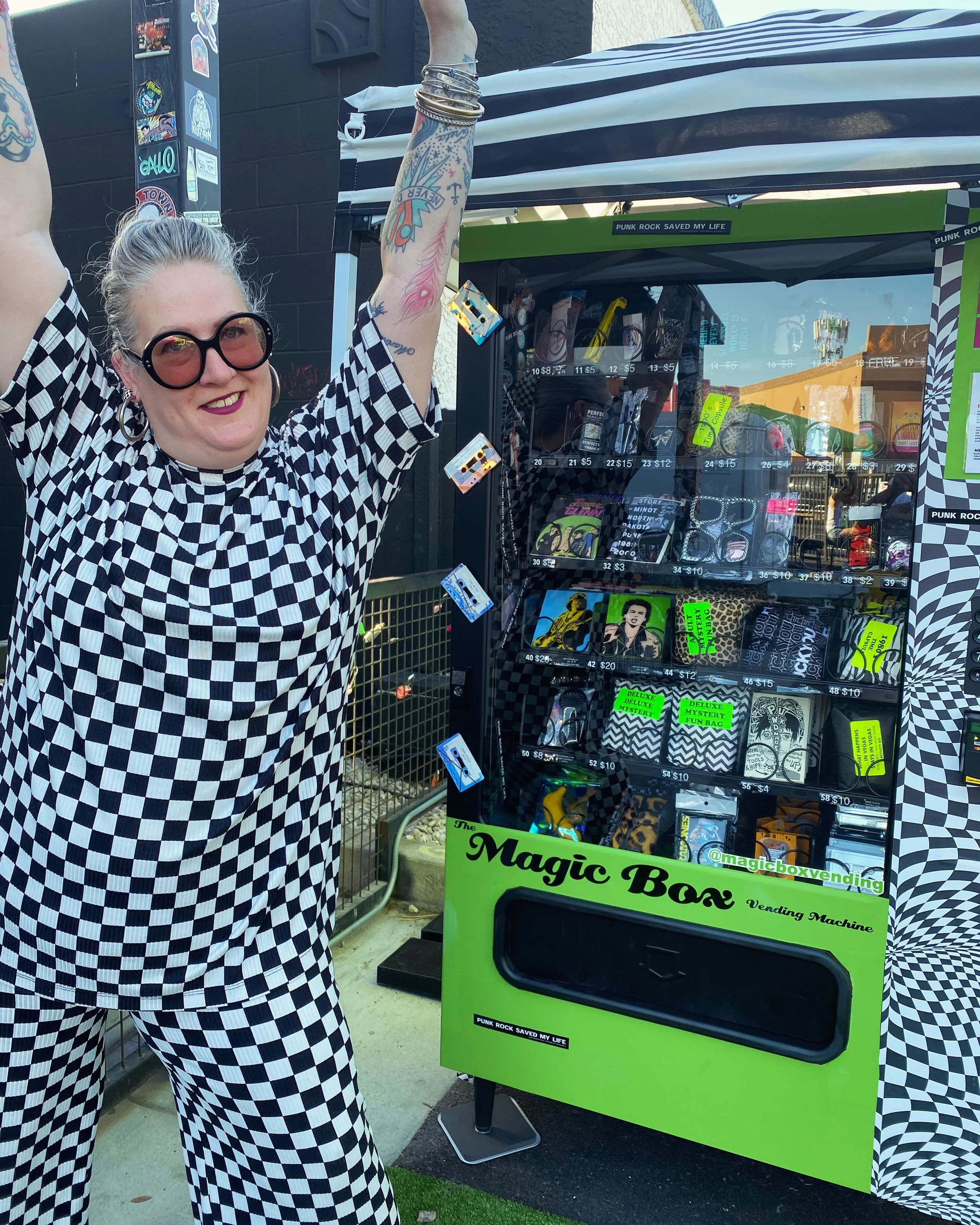 A picture of an enthusiastic blonde woman with her arms raised in celebration. She is wearing large, stylish sunglasses and a black and white checkerboard two piece set. She is standing in front of a magic box vending machine.