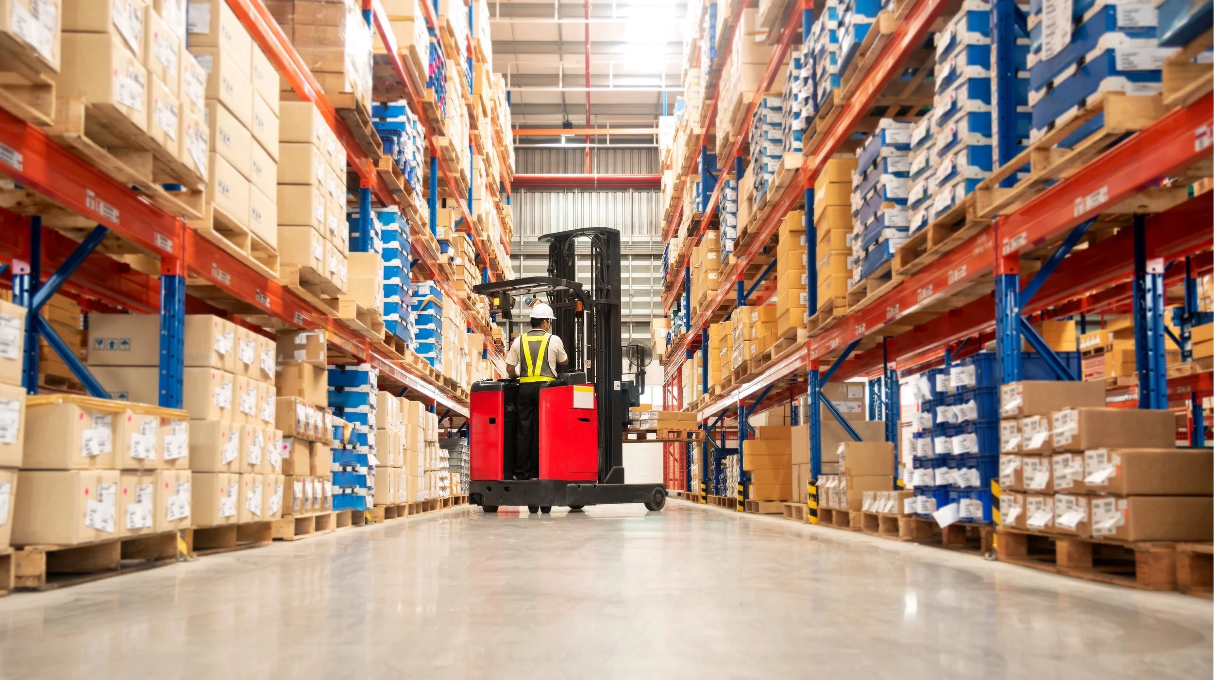 A warehouse worker operating a forklift among tall shelves filled with boxes and pallets.