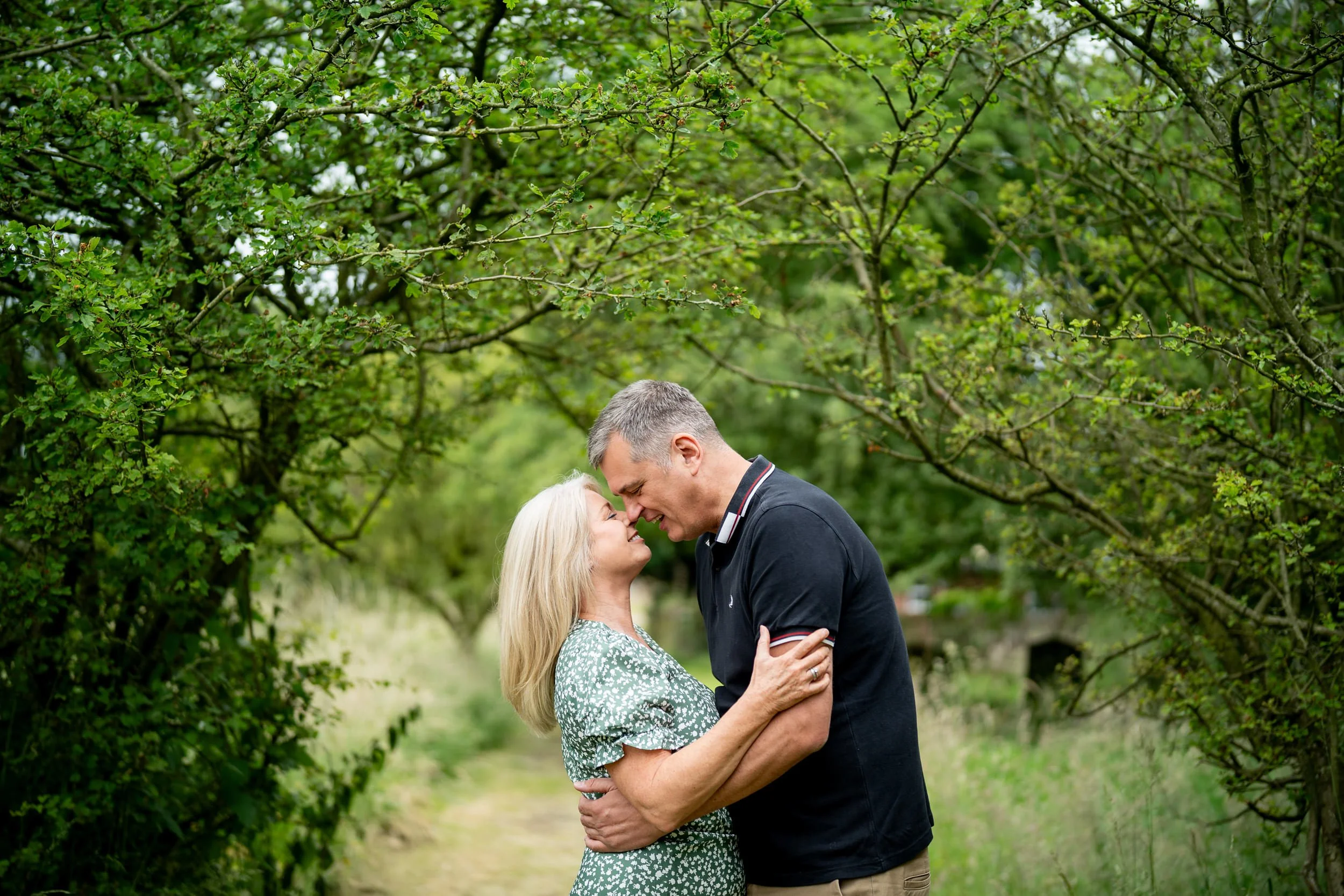 A middle-aged woman and a middle-aged man stand close together on a nature trail, smiling and touching foreheads amidst green foliage and trees.