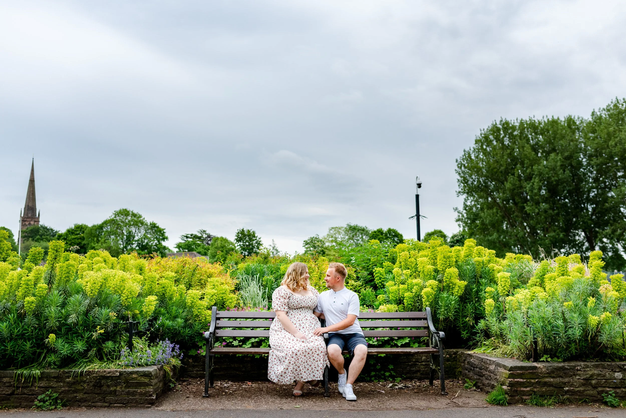 A man and woman sit on a park bench surrounded by lush green plants and flowers, looking at each other, with a cloudy sky and trees in the background.