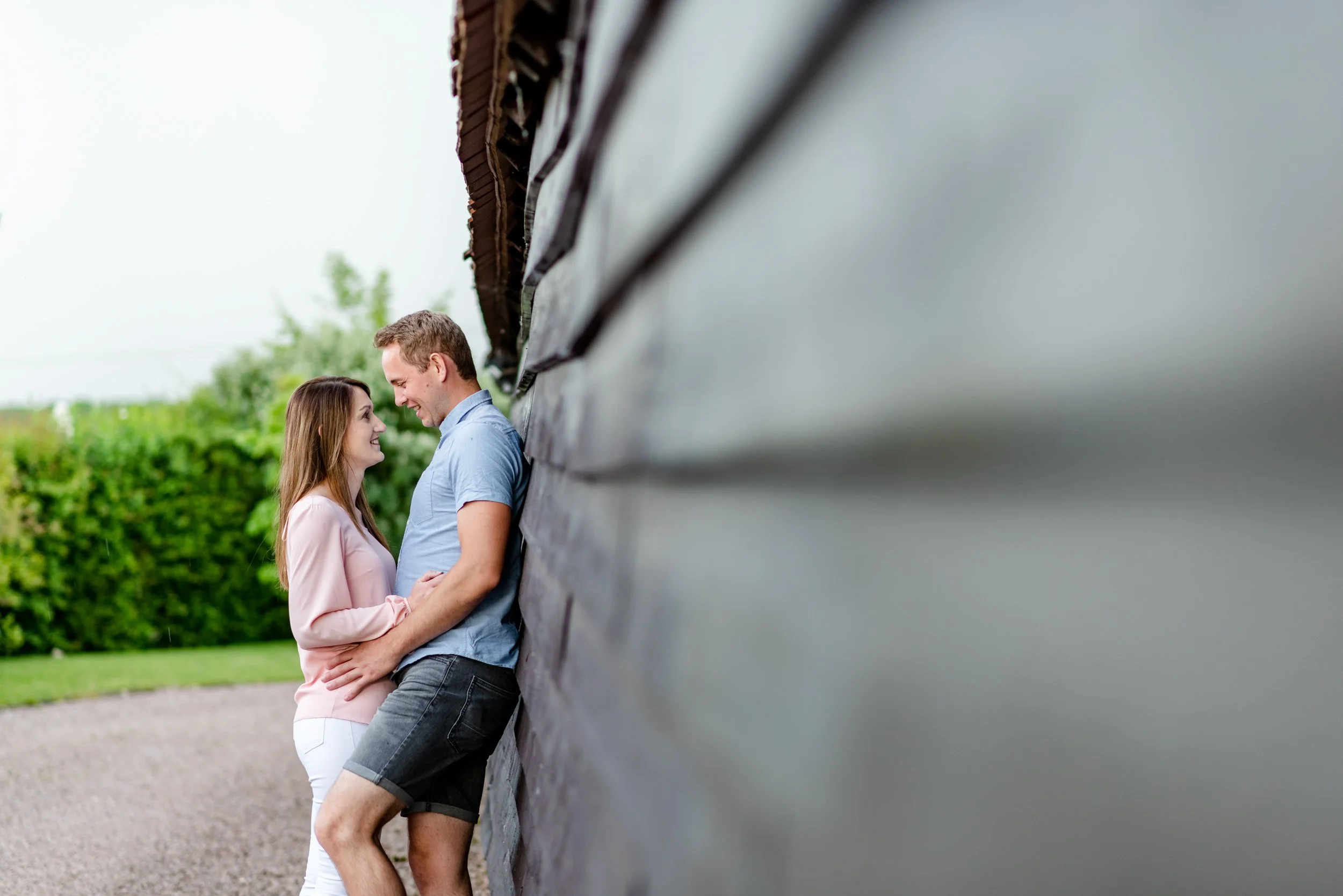 A couple standing close, smiling and looking into each other's eyes, leaning against a dark wooden wall outdoors with green bushes in the background.