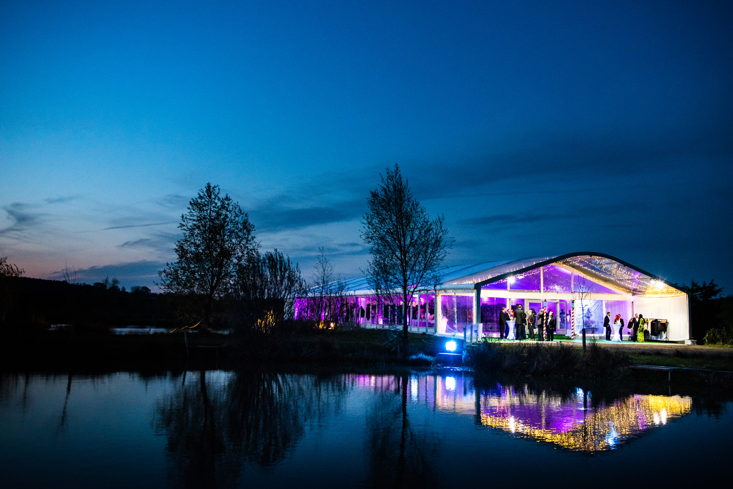 Evening shot of a Fews Marquee colourfully lit up against the night sky and reflected in a pond