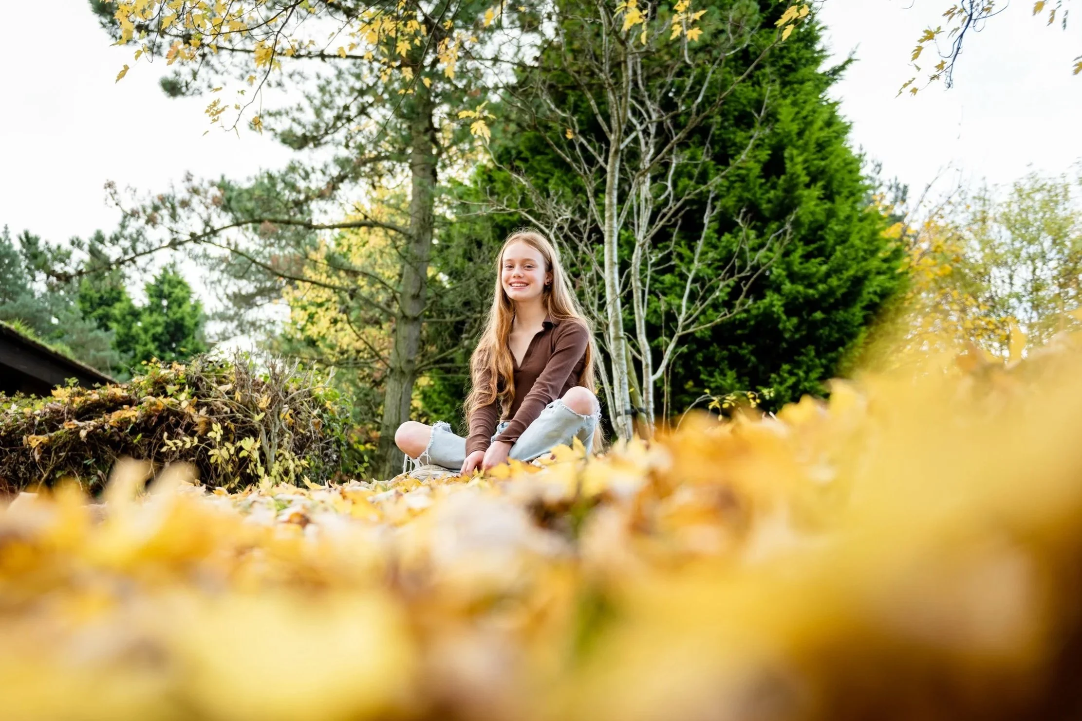 A young girl with long hair sitting cross-legged outdoors in a park during fall, surrounded by fallen yellow leaves and trees with autumn foliage.