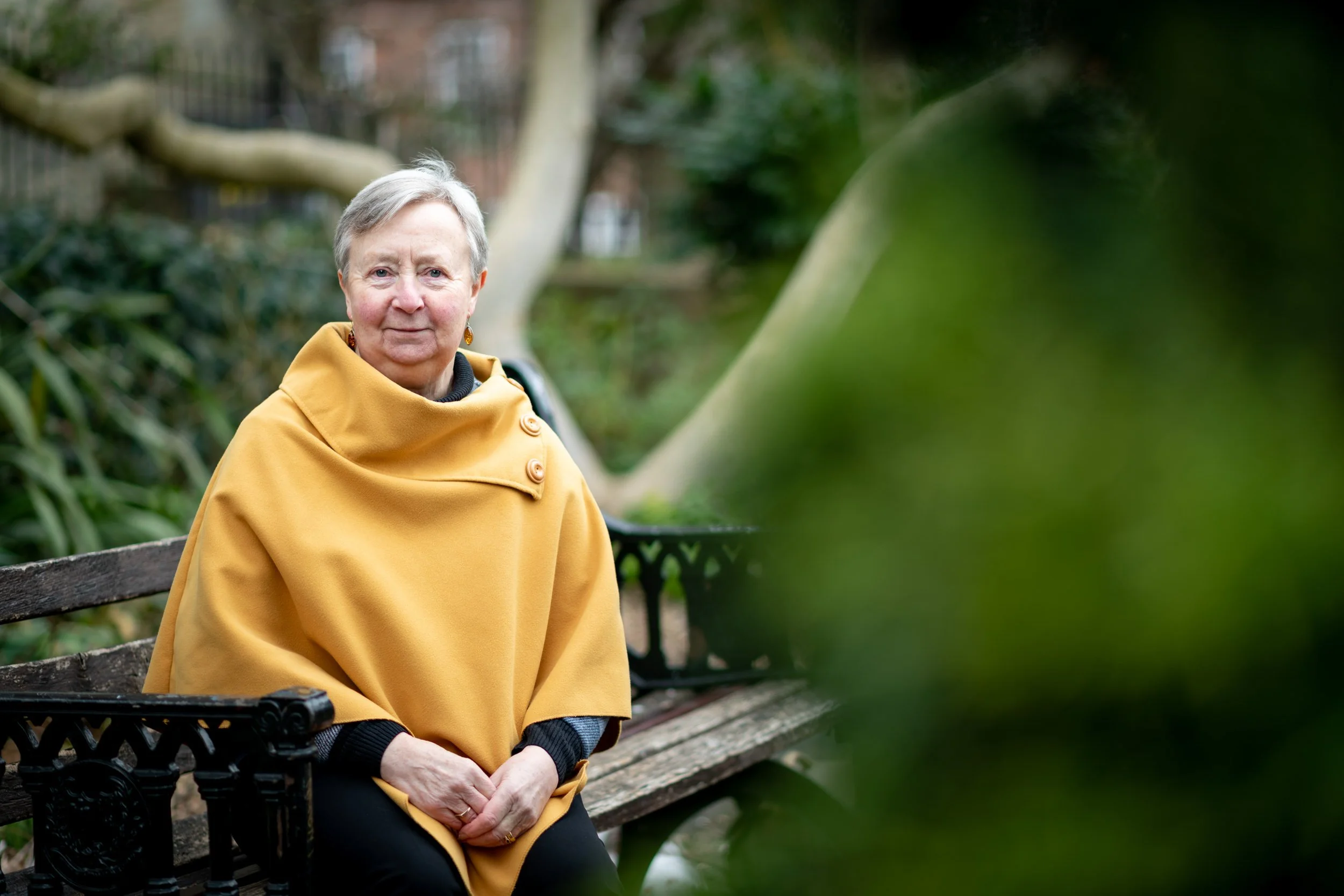 An elderly woman with short gray hair sitting on a park bench wearing a yellow poncho and black pants, with greenery and trees in the background.