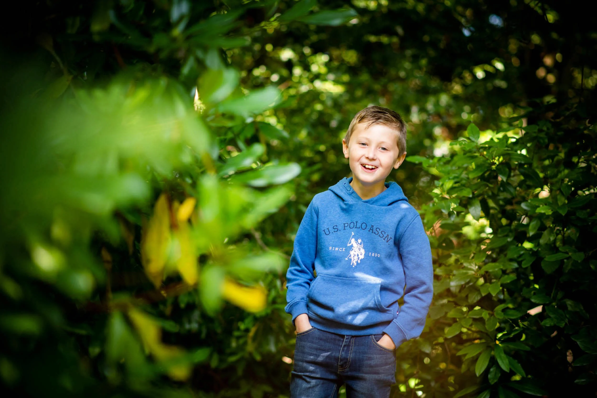 A young boy in a blue hoodie standing in a lush green garden, smiling and looking happy.