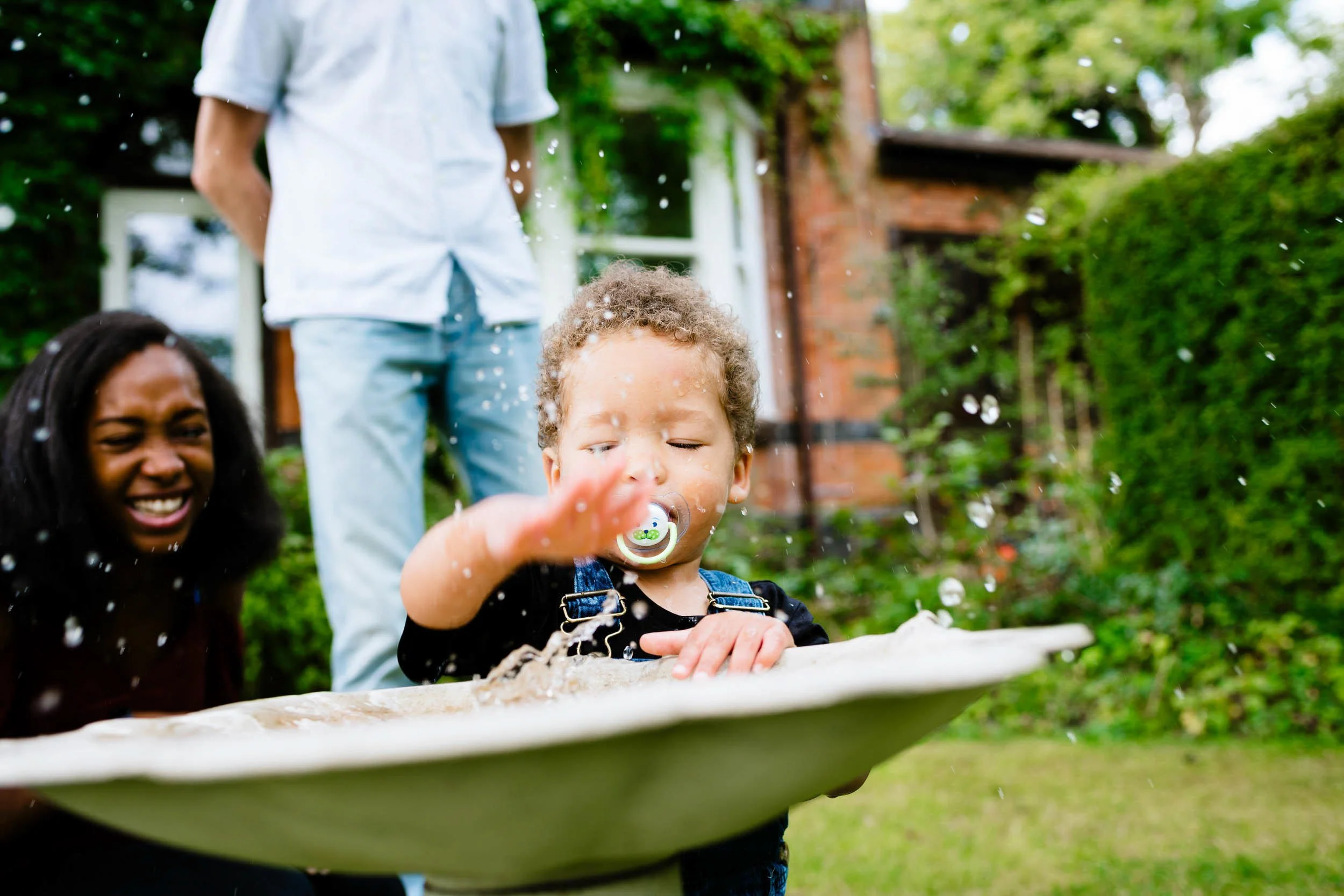 A young boy with curly hair and a pacifier in his mouth is reaching out to splash water from a basin. An adult woman with dark curly hair is smiling, and a man is standing nearby in a garden with lush green trees and shrubs.