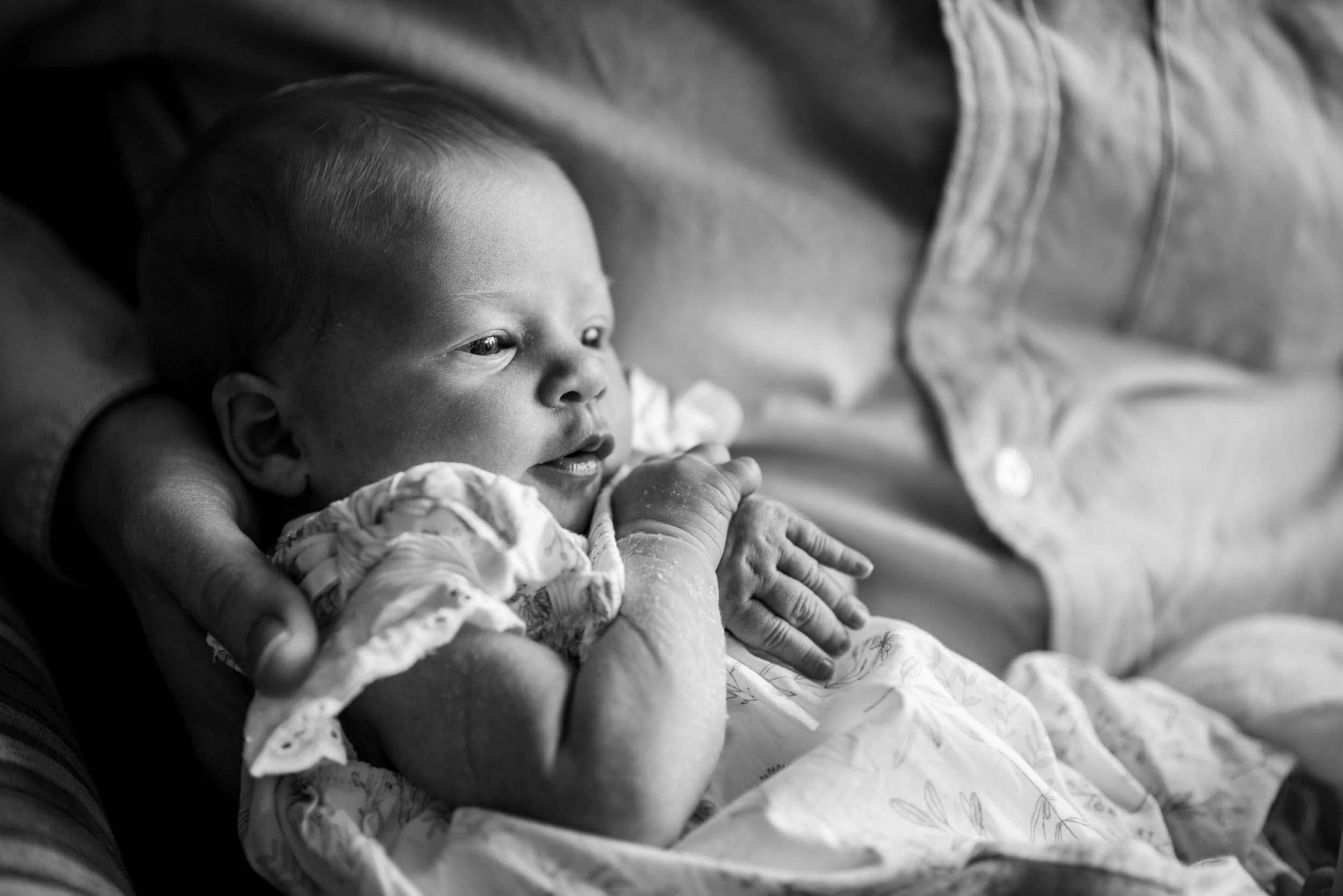 Black and white photo of a baby in a crib, lying on its stomach, with a person’s arm around him, wearing a short-sleeved patterned outfit.