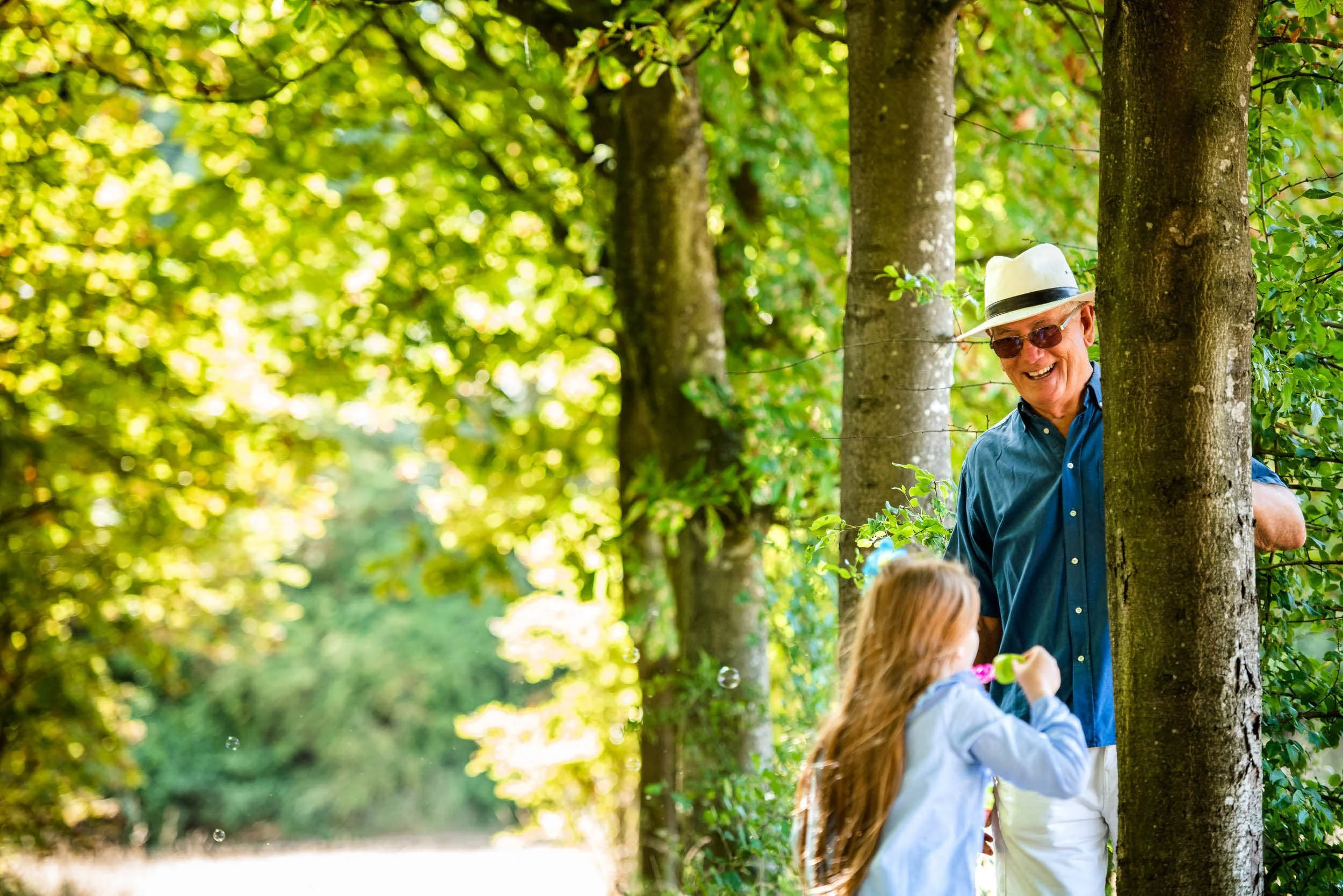 An elderly man wearing a white hat, sunglasses, a blue shirt, and white pants is smiling while peeking from behind a tree. A young girl with long hair, wearing a light-colored shirt, is playing with bubbles in a lush green park.