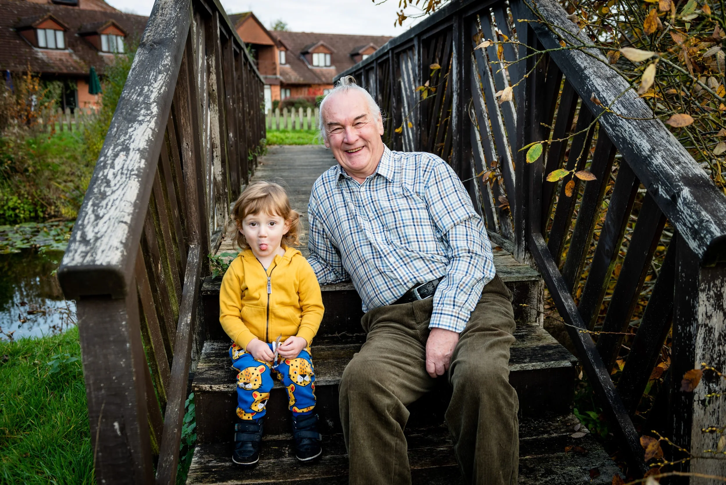 An elderly man and a young girl sitting on a wooden bridge outdoors. The man is smiling widely, wearing a plaid shirt and brown corduroy pants. The girl, in a yellow jacket and patterned pants, is making a funny face. In the background, there are hou