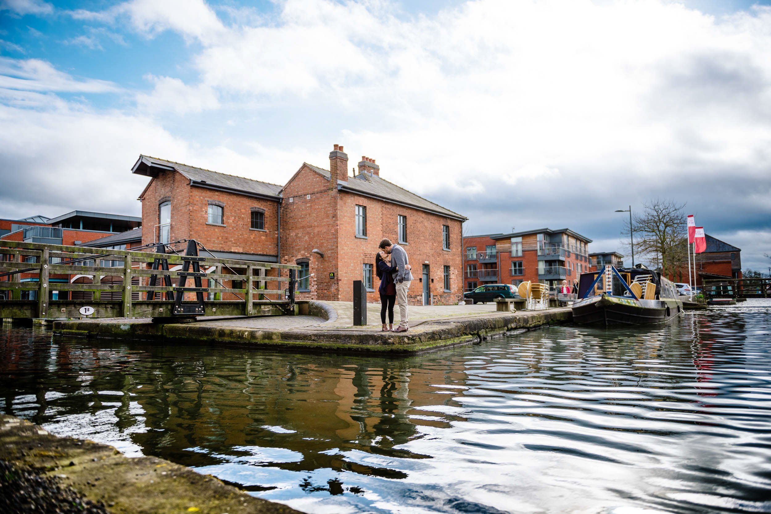 A couple standing on a canal bank, embracing and kissing with a brick building in the background, boats on the water, and modern apartment buildings nearby under a cloudy sky.
