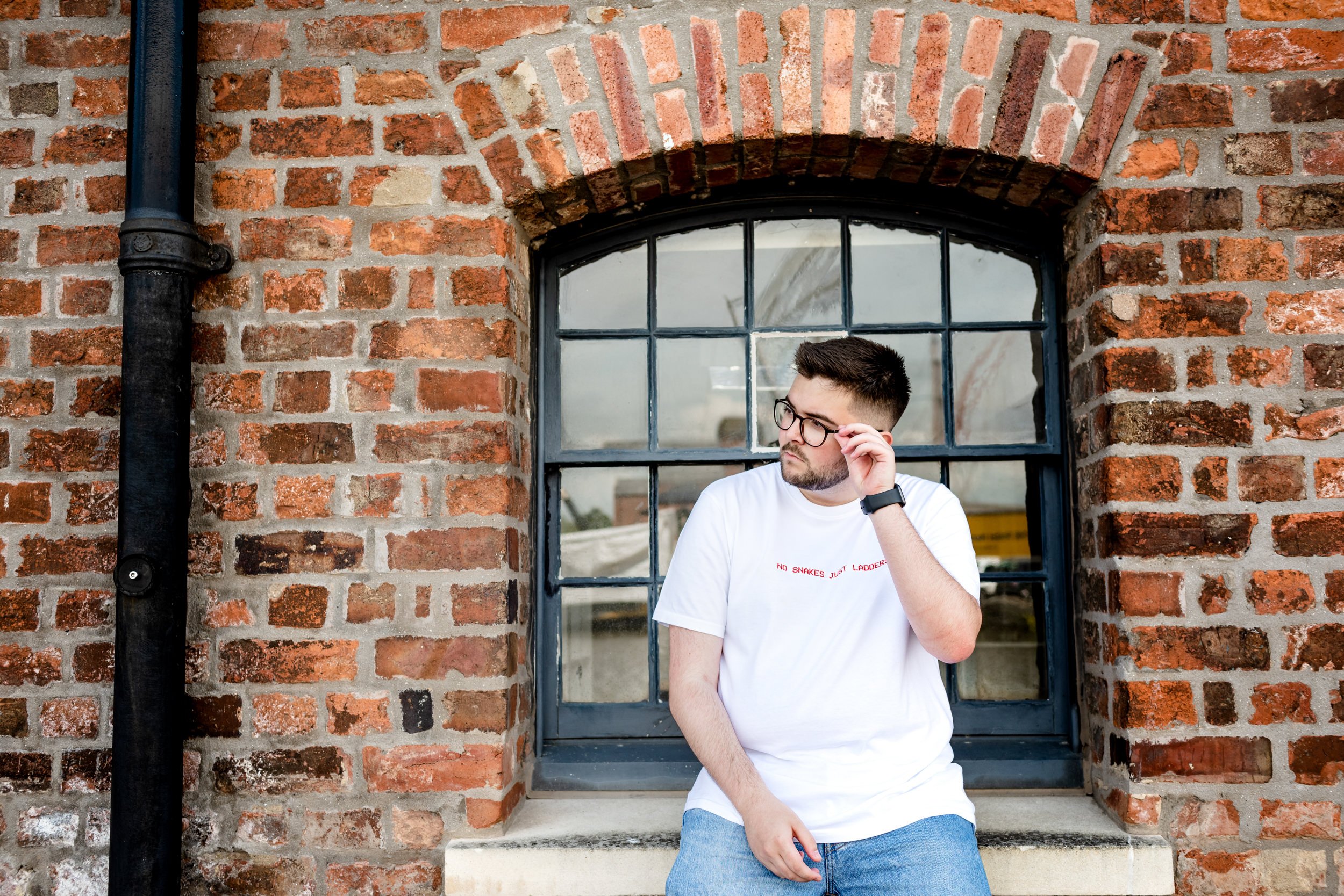 A young man with dark hair and glasses, wearing a white T-shirt and jeans, sitting on a concrete window ledge, adjusting his glasses, against an exposed brick wall.