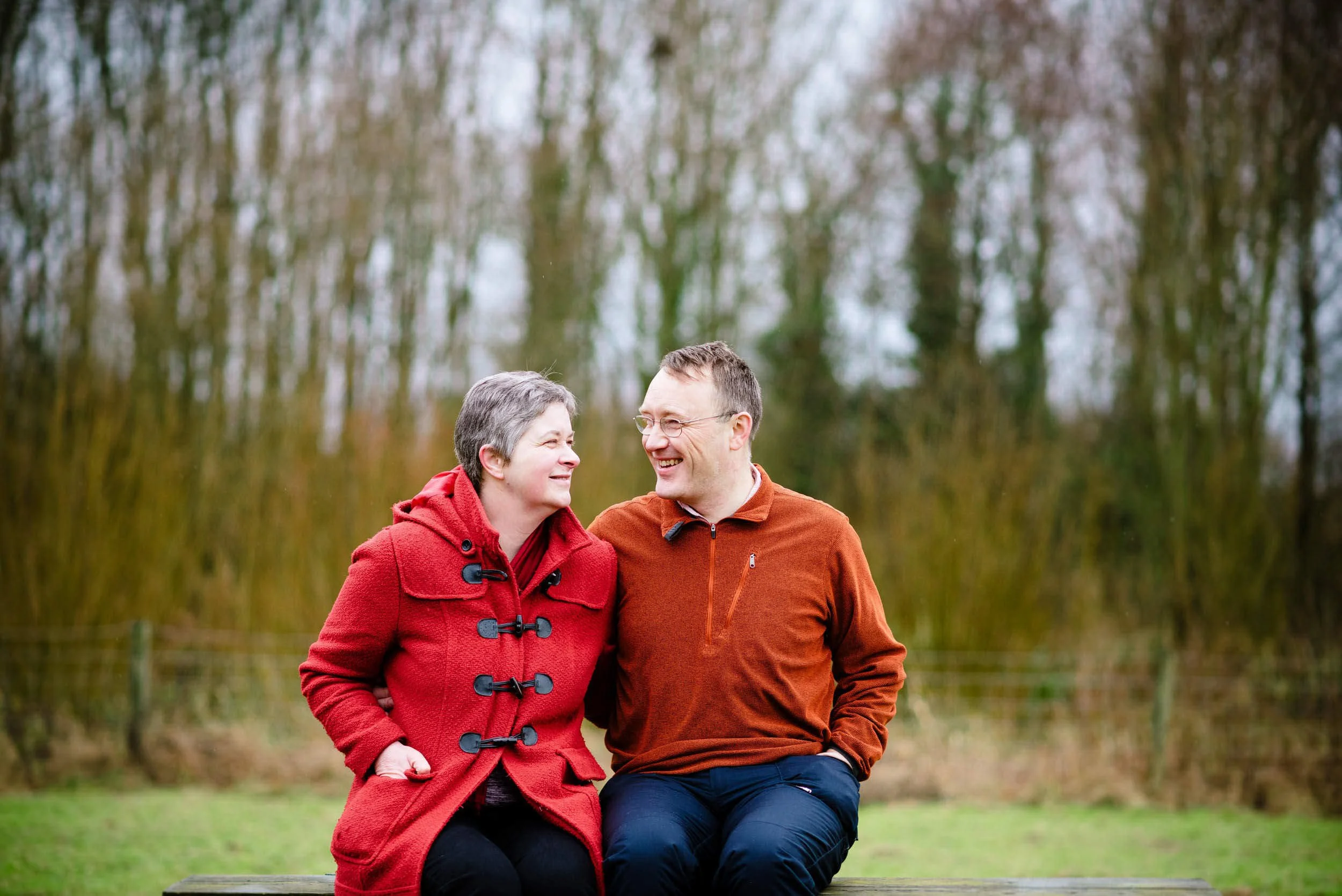 A man and woman sitting on a bench outdoors, smiling and looking at each other, with trees in the background.