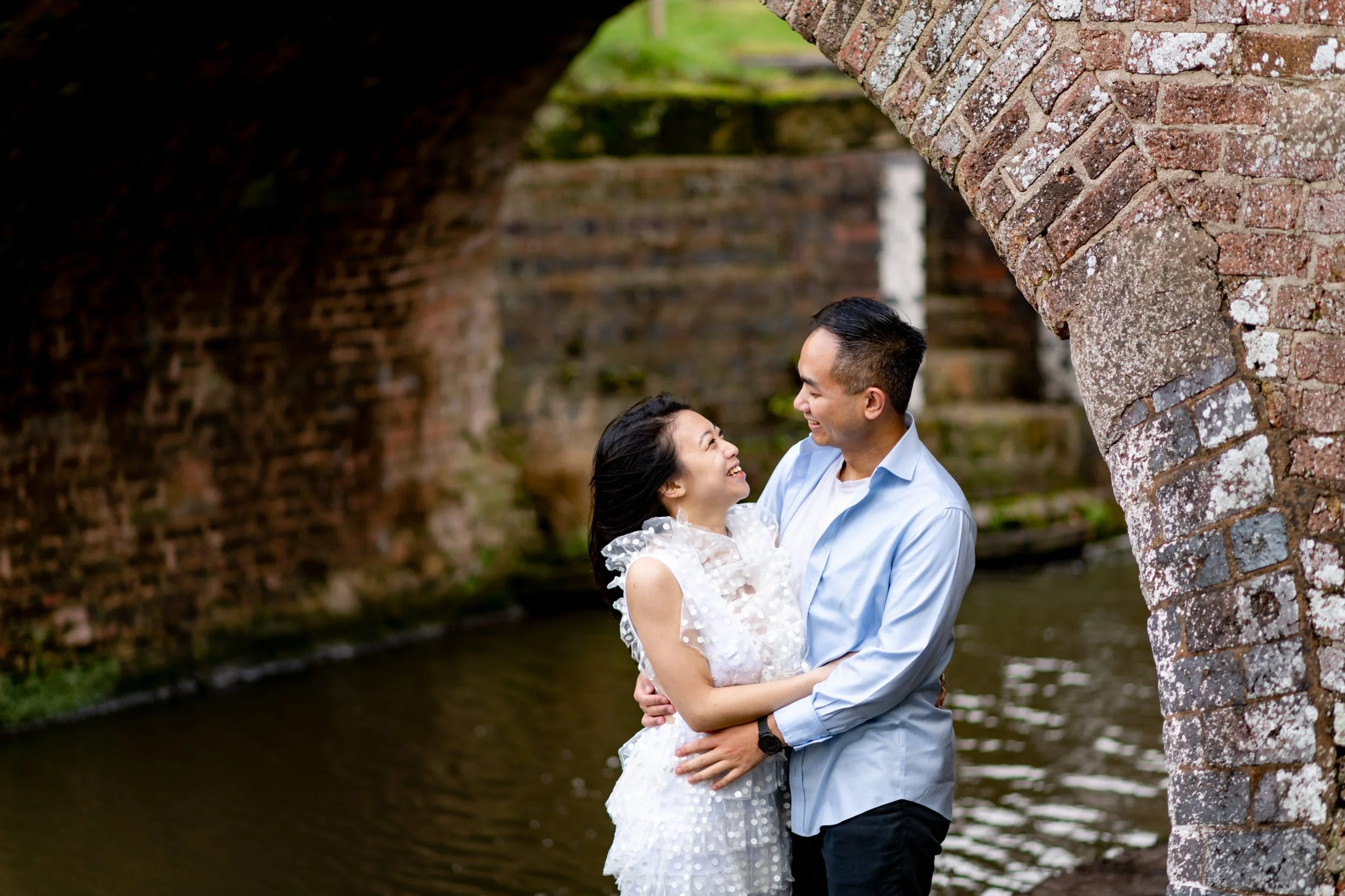 A joyful couple standing close together under a brick bridge near water, smiling and looking at each other, with the woman wearing a white dress and the man in a light blue shirt.