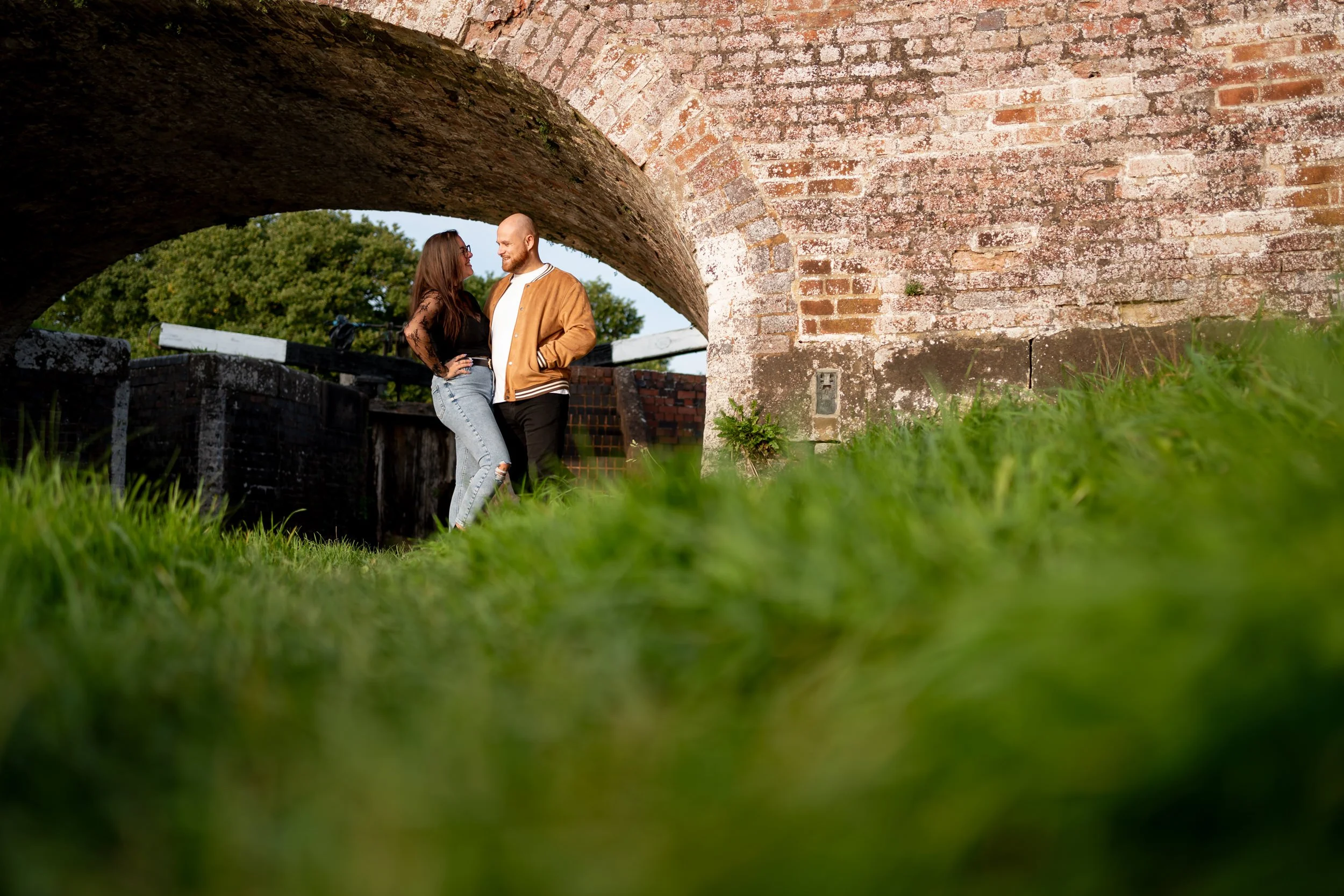 A couple standing close together, smiling and looking at each other, under a stone bridge with green trees in the background, viewed from a low angle on the grass.