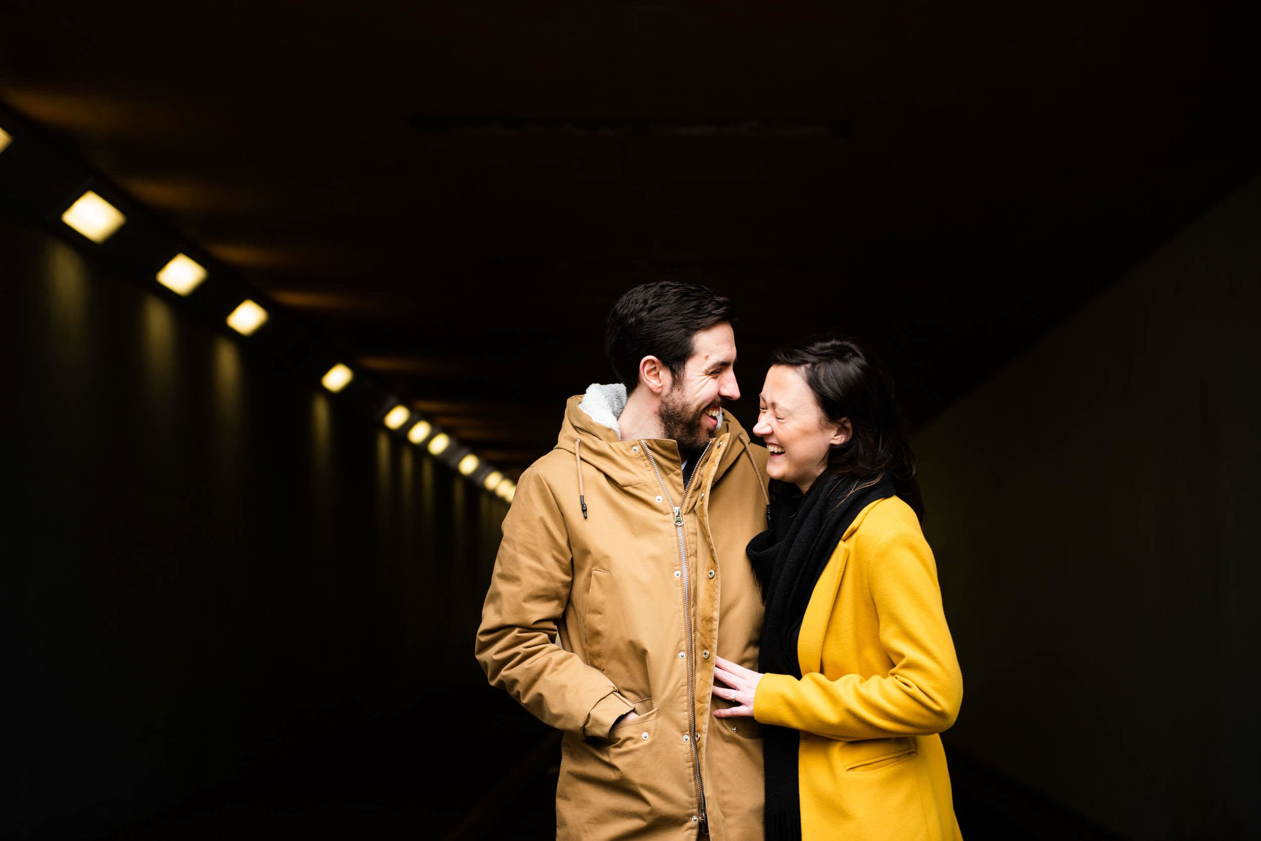 A smiling couple standing close together in a dark tunnel, wearing yellow and tan winter coats.