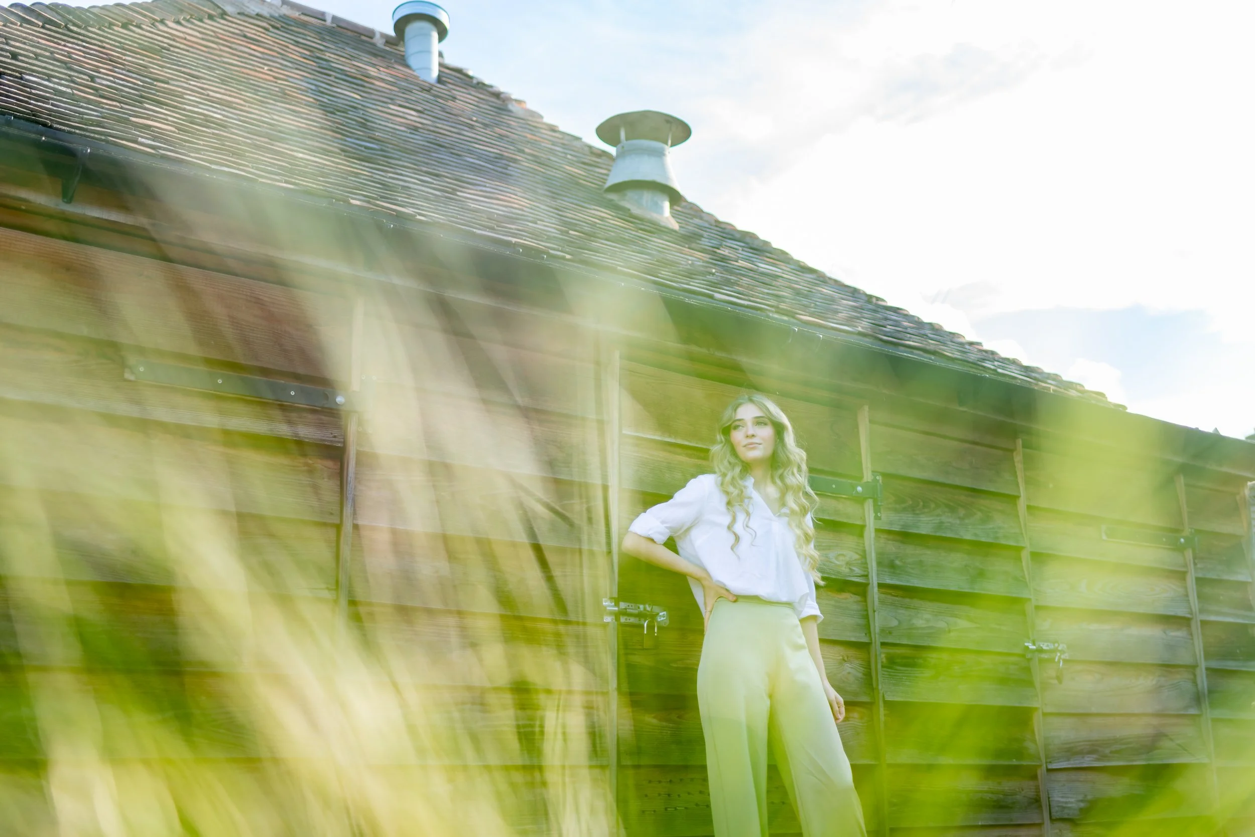 A woman with long, wavy blonde hair wearing a white blouse and beige pants standing outside in front of a wooden fence and a house with a tiled roof, with greenery in the foreground and a partly cloudy sky in the background.