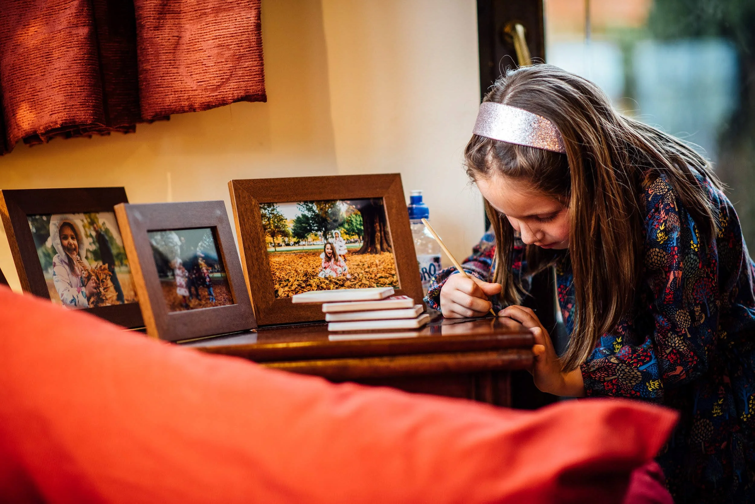 A girl with long brown hair and a sparkly headband is leaning over a table, playing a game or doing an activity with a small stick. On the table, there are three picture frames displaying photos of a young girl outdoors in autumn, smiling and sitting